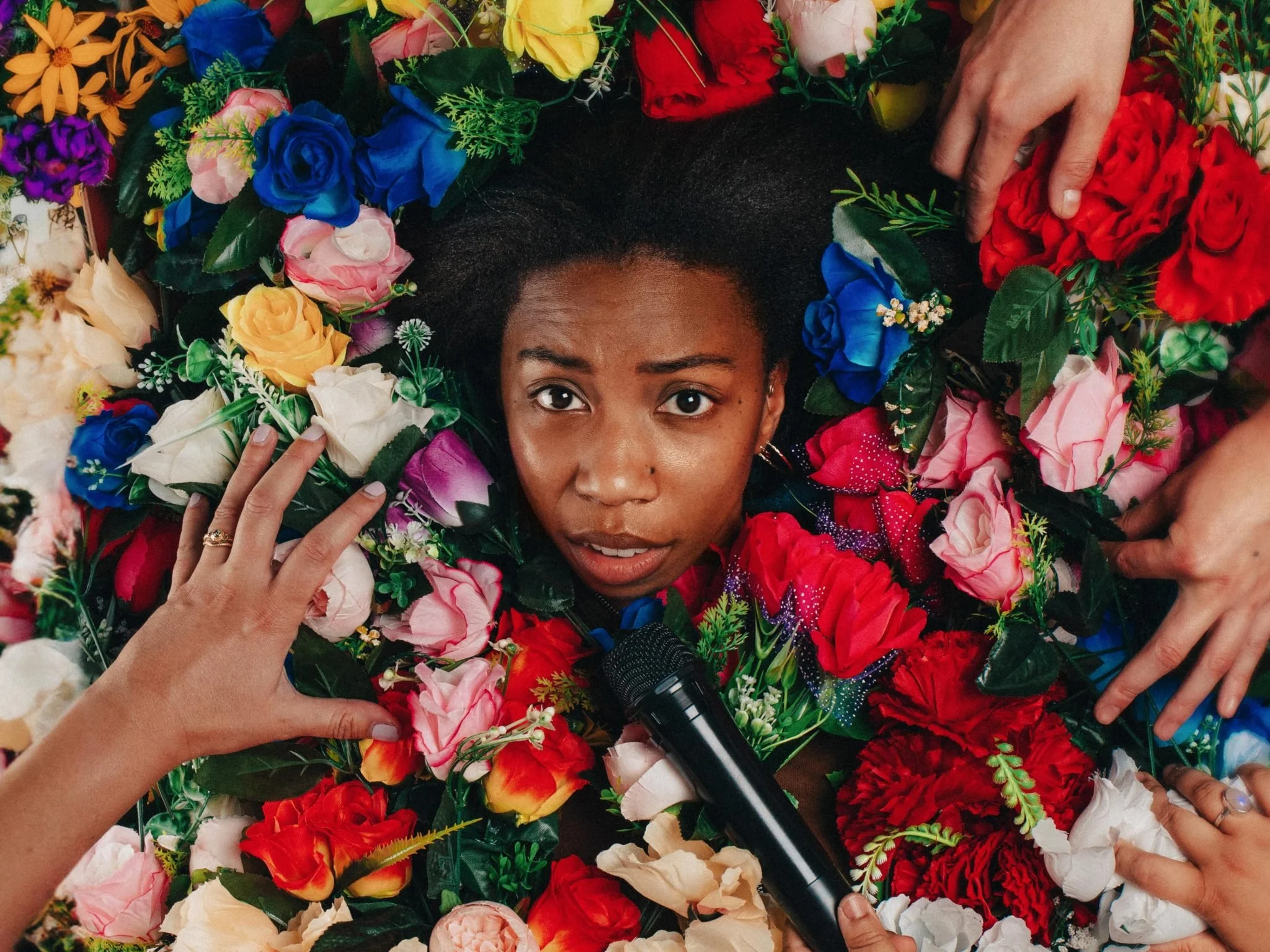 A woman lying on the floor surrounded by colorful artificial flowers, holding a microphone.