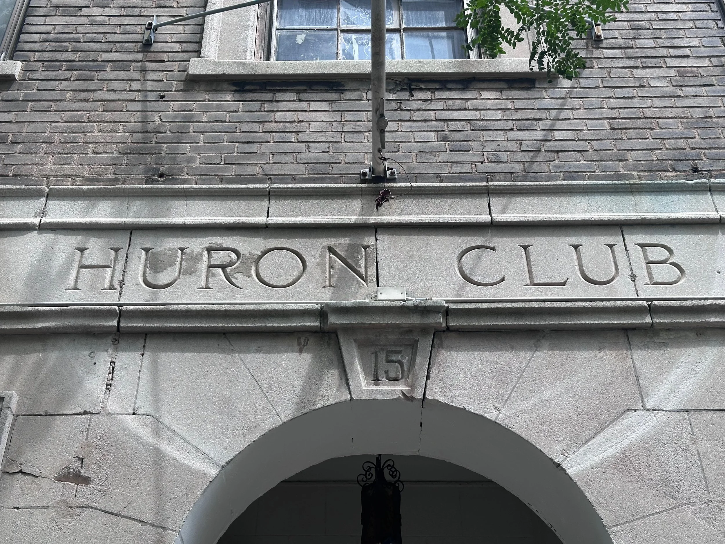 Close-up of a building's exterior showing gray brick and stonework with the engraved words 'Huron Club' on the stone arch beneath a window with a metal railing and greenery.