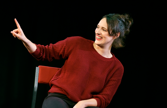 A young woman with dark hair in a bun, wearing a red sweater, smiling and pointing her finger to the left on a black background.