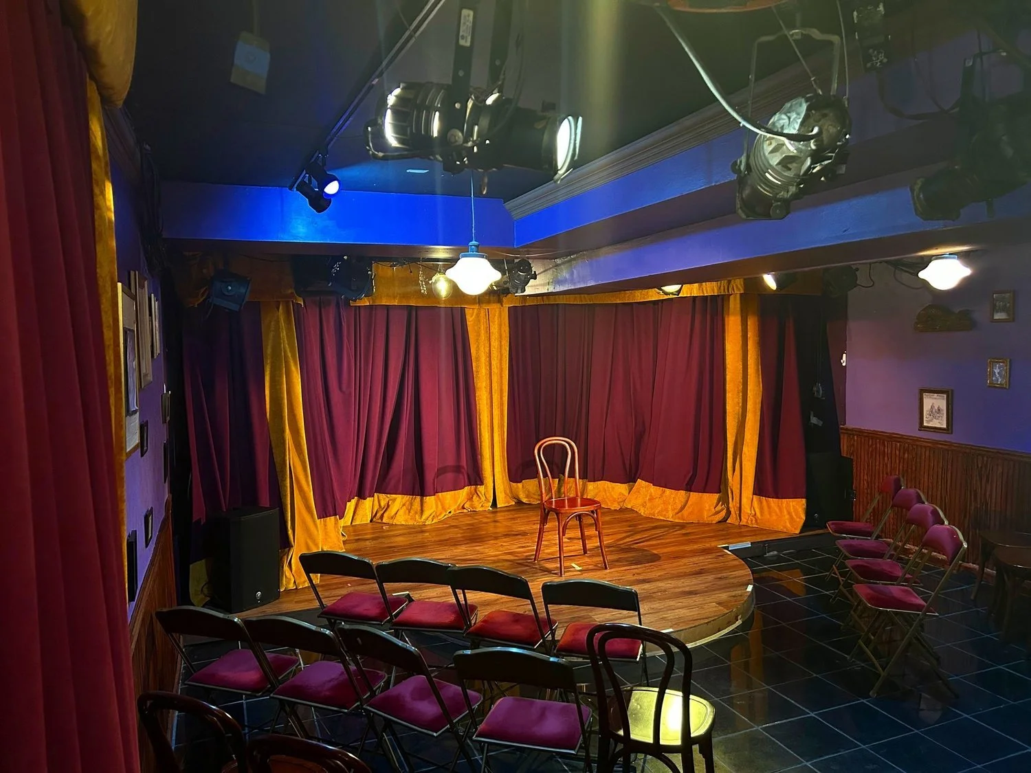 Small theater stage with a wooden floor, surrounded by purple curtains with gold trim, and a single wooden chair at center stage. Seating chairs facing the stage are arranged in rows. Overhead stage lights are directed at the stage.