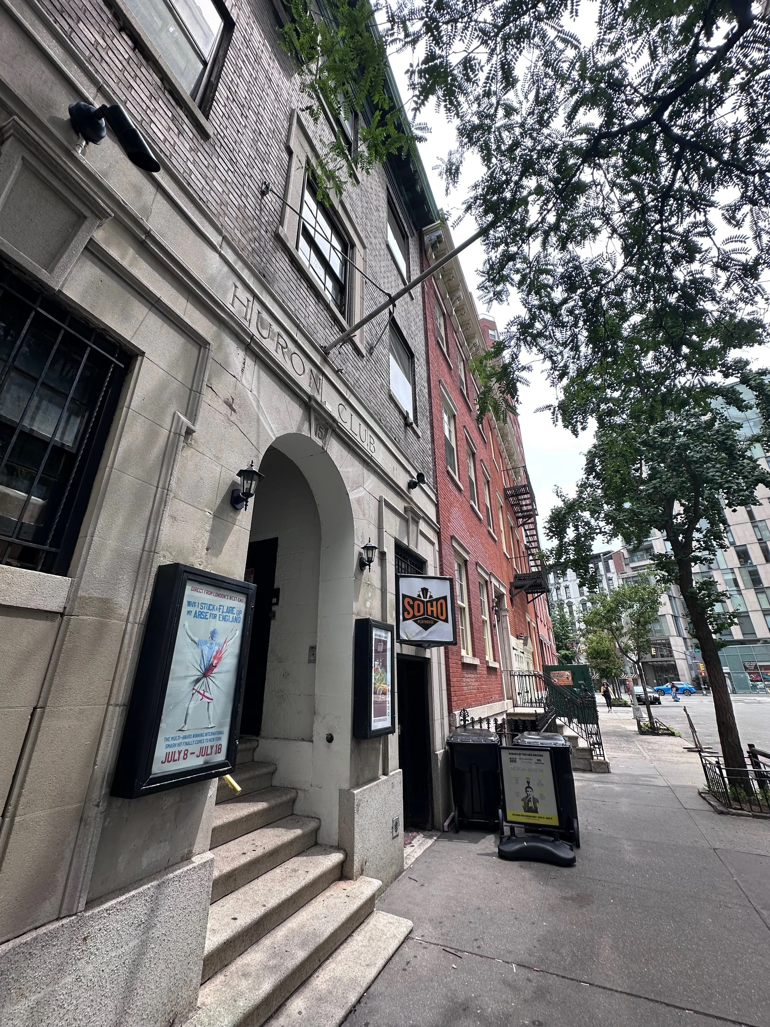 Street view showing a brick building with signs for a theater and other businesses, trees, and sidewalk sidewalk in an urban area.