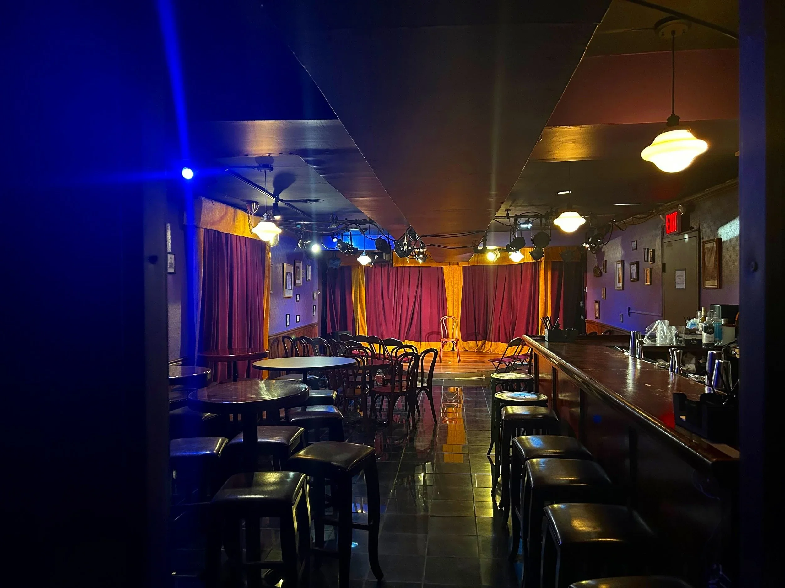 Empty stage with red curtains and row of chairs, illuminated by warm lighting, located inside a performance venue or comedy club.