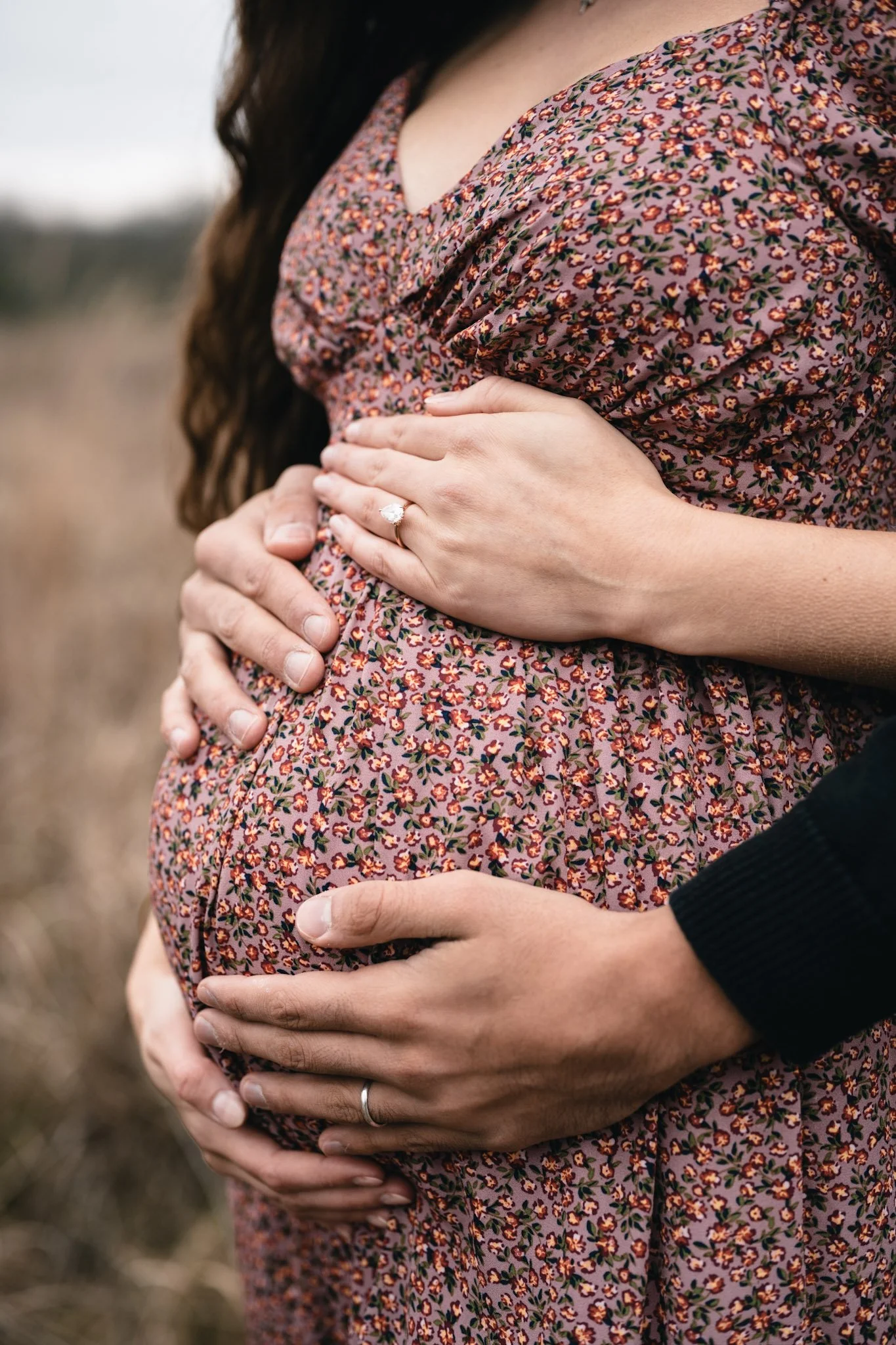 Close-up of a pregnant woman wearing a floral dress with a man’s hands gently cradling her belly from both sides, one hand displaying a wedding ring, outdoors with blurred background.