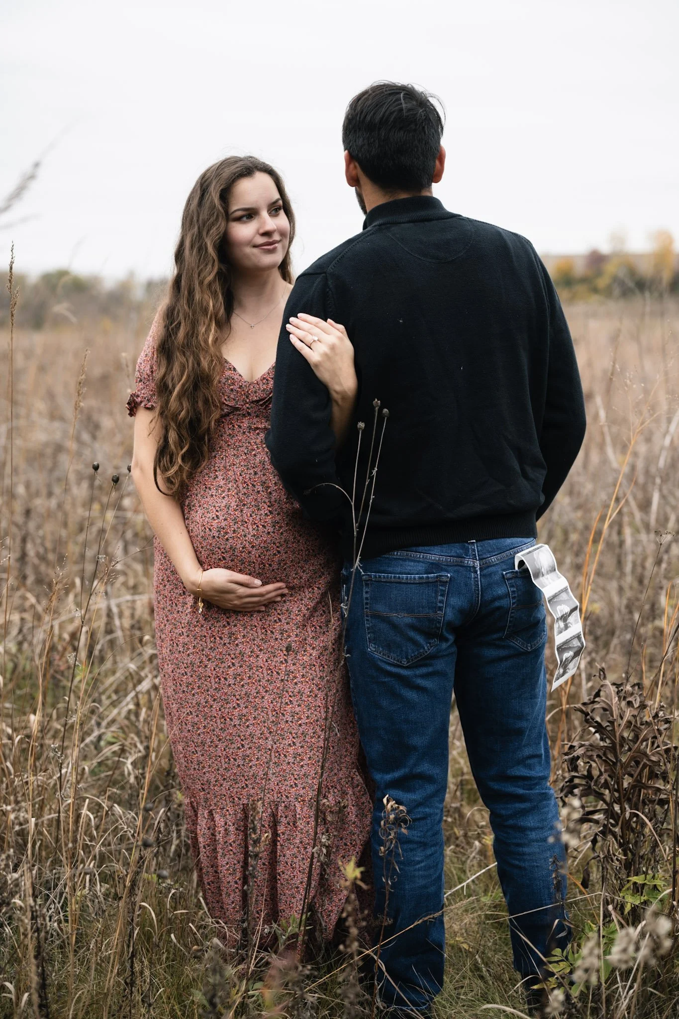A pregnant woman with long wavy hair in a floral dress standing in a field, holding her belly, as a man with short dark hair and a beard in a black jacket and jeans faces her. She gently touches his shoulder with her hand. The sky is overcast and the field has dry grasses and plants.