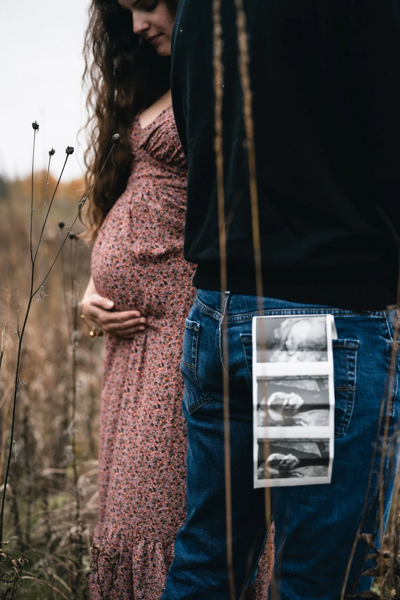 A pregnant woman in a floral dress stands outdoors with her hand on her belly, next to a man in jeans and a dark jacket, who is holding ultrasound images on his pocket.