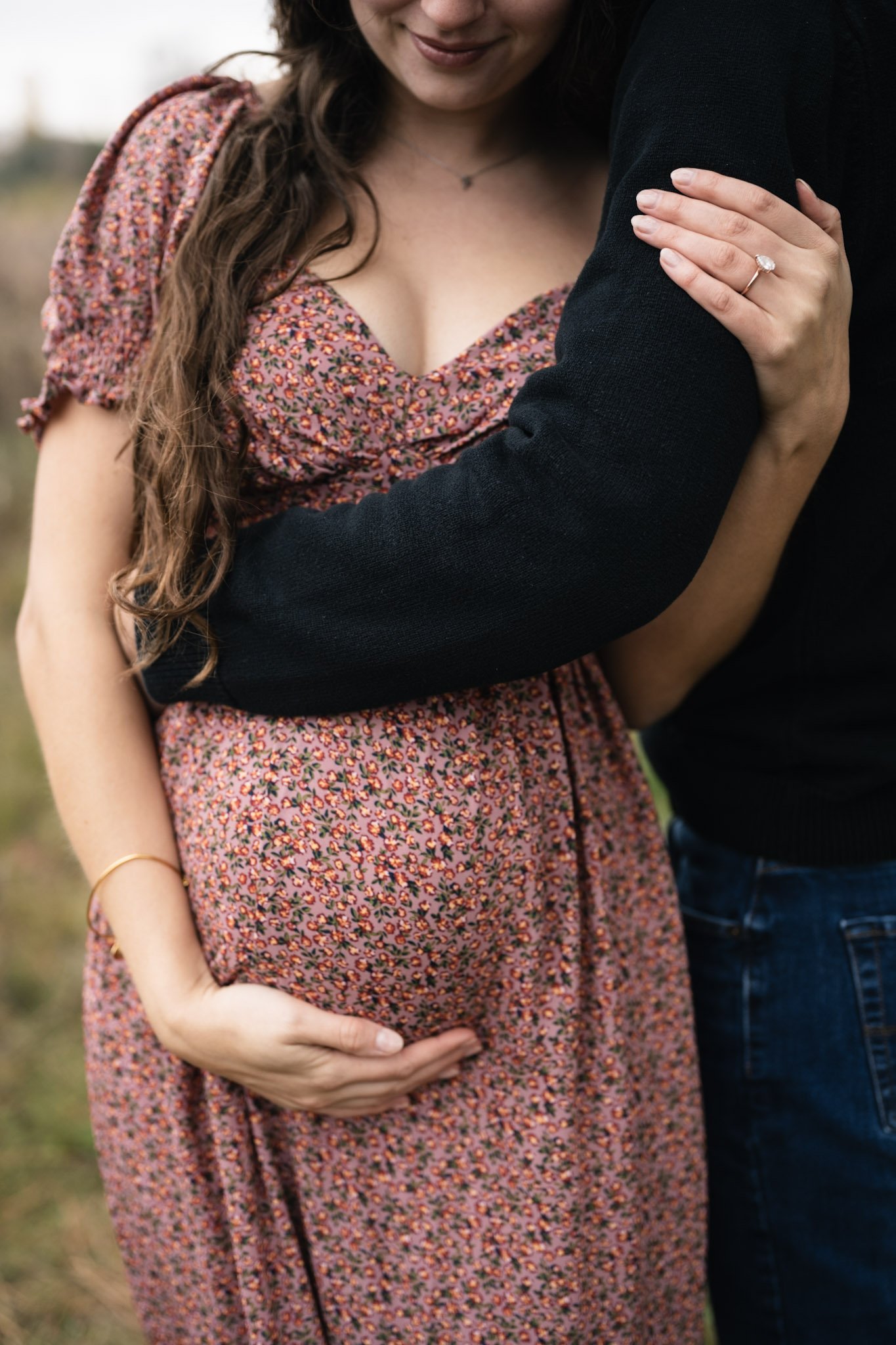 A pregnant woman in a floral dress resting her hand on her belly while she is being embraced by a man wearing a black sweater. The woman has long, wavy brown hair and is wearing a ring on her left hand.