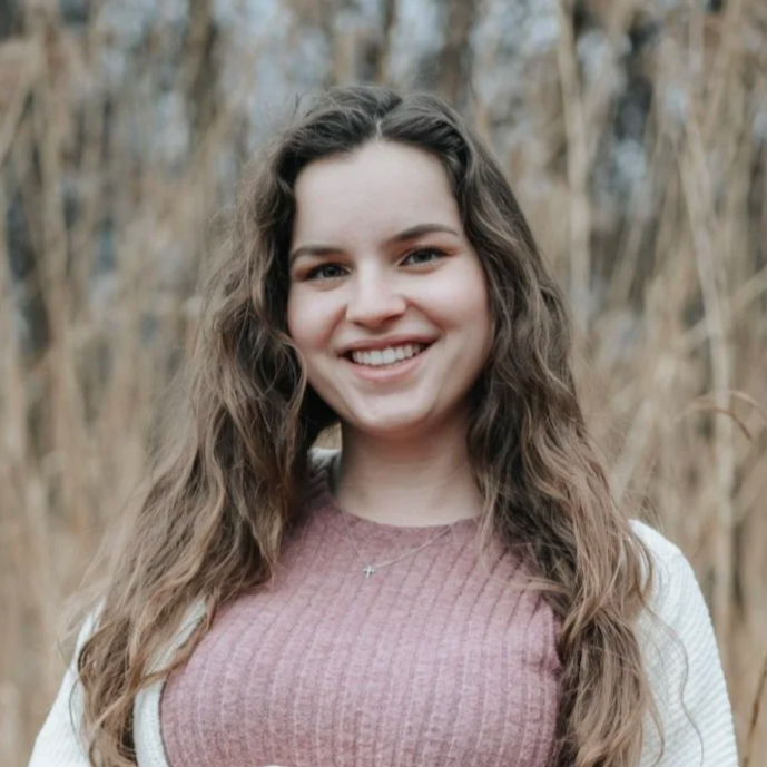 A young woman with long, wavy brown hair smiling outdoors in front of a background of dry, leafless branches.
