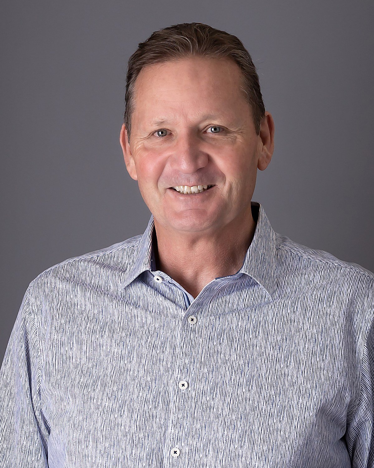 Headshot of a smiling middle-aged man with short brown hair, wearing a light blue striped collared shirt against a gray background.
