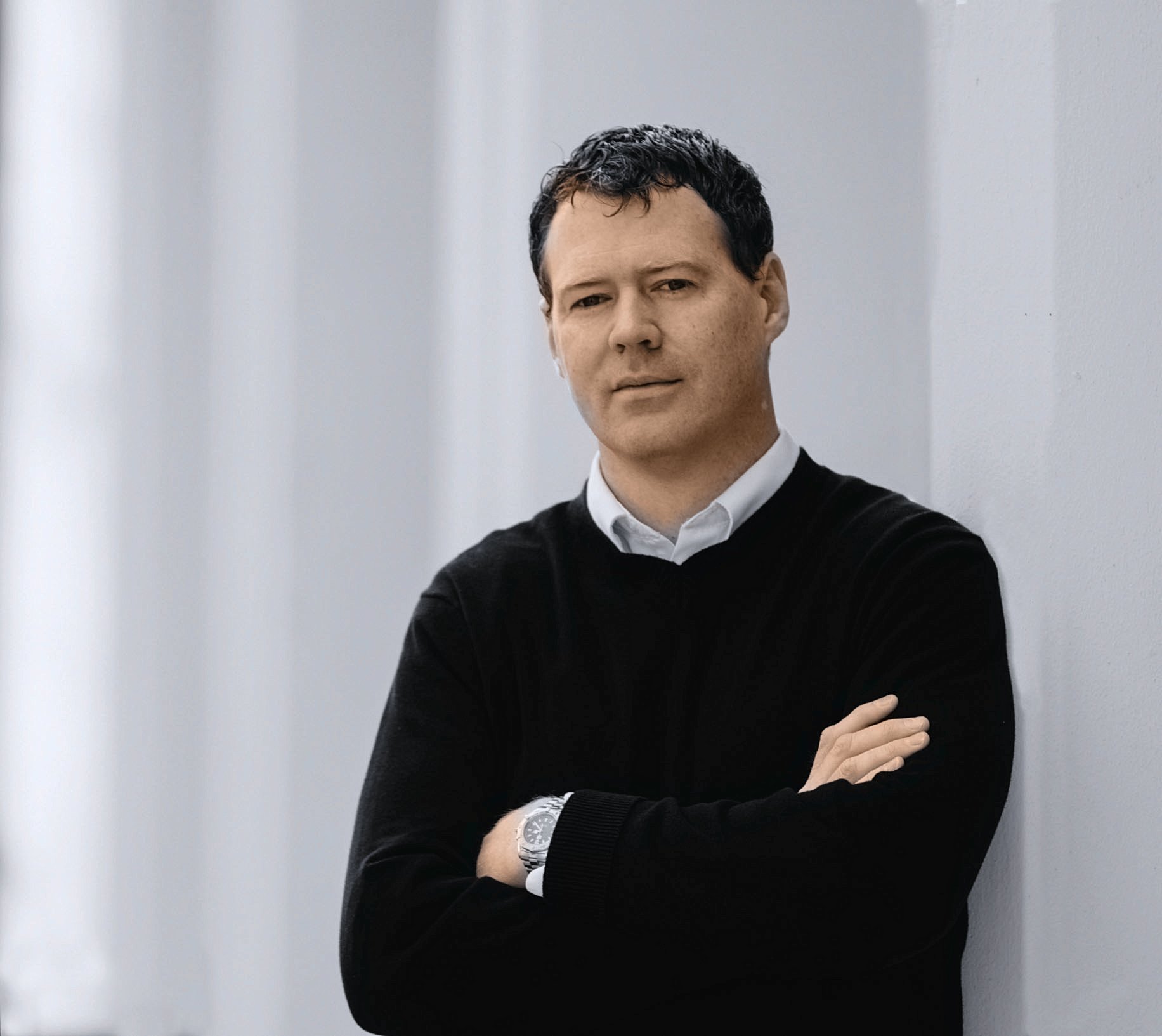 A man with dark, curly hair and light skin, dressed in a black sweater over a white shirt, standing in front of a plain light-colored wall, with arms crossed, looking at the camera.