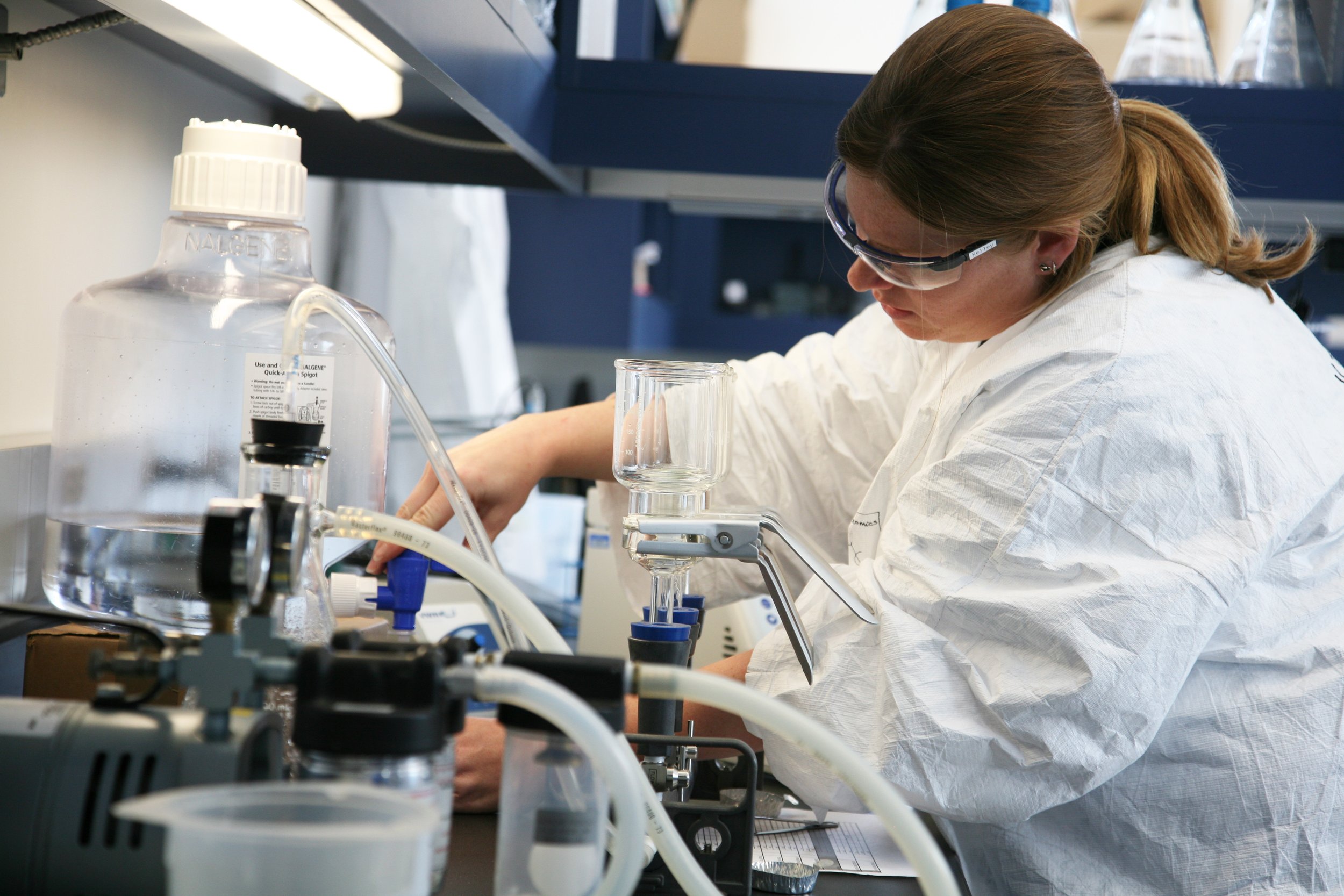 A scientist wearing safety glasses and a white lab coat working with laboratory equipment and glass containers in a laboratory setting.