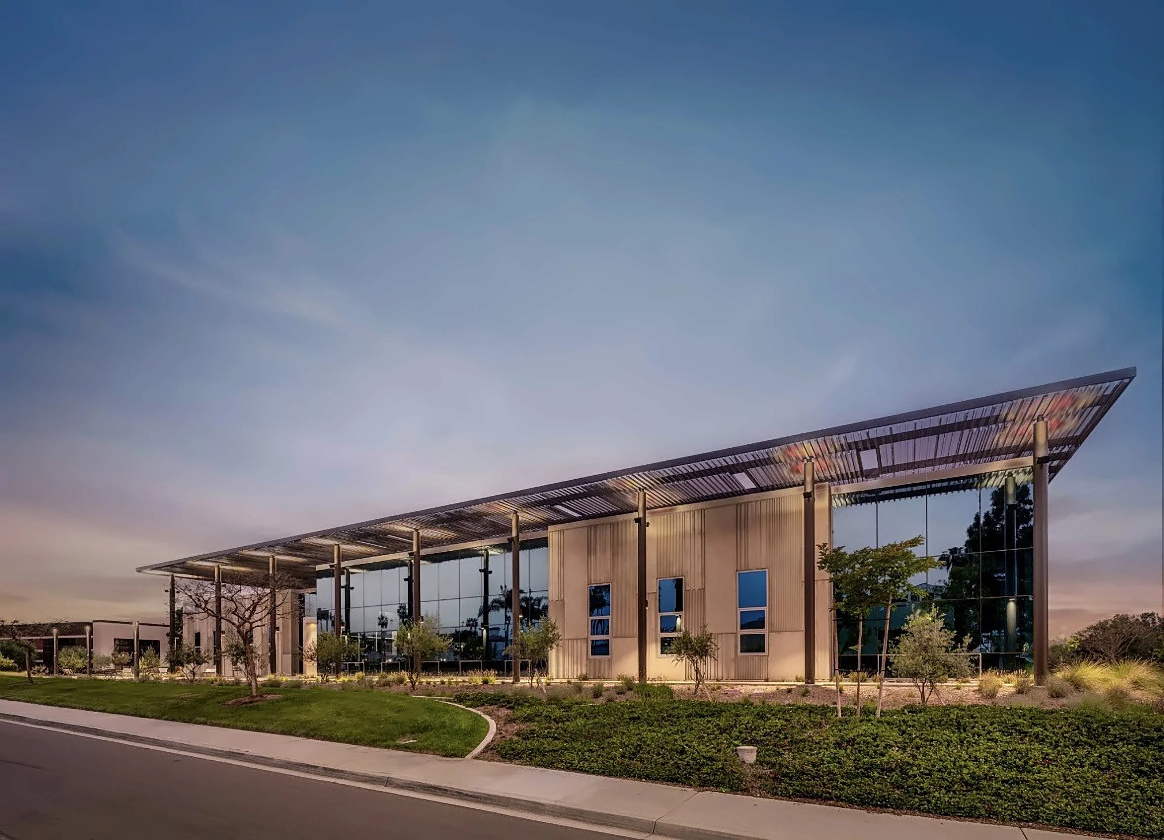 Modern building with large glass windows and a metal overhang, surrounded by landscaped trees and grass, under a clear sky at dusk.