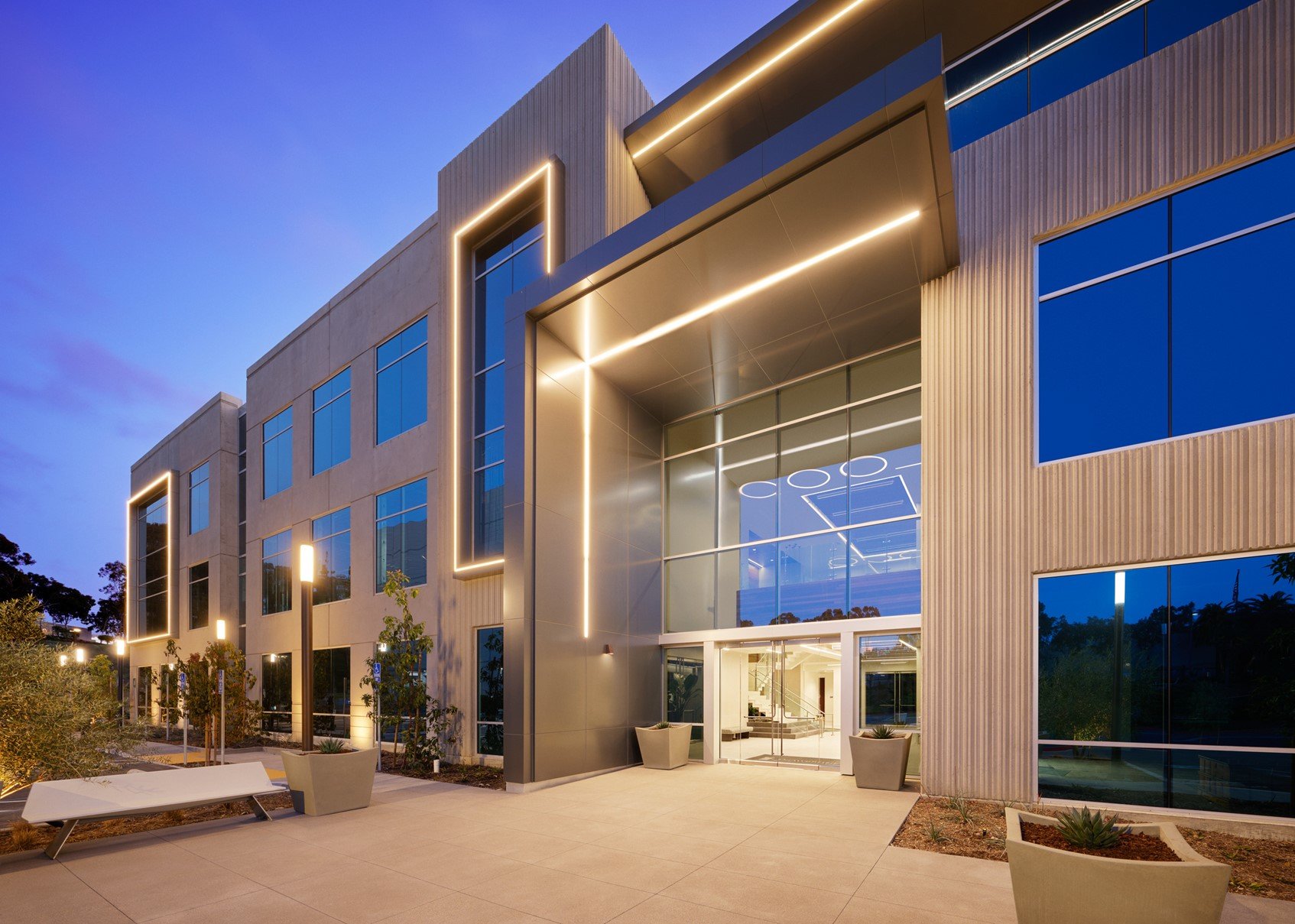Modern commercial building at dusk with large glass facade, illuminated trim, and landscaped exterior.