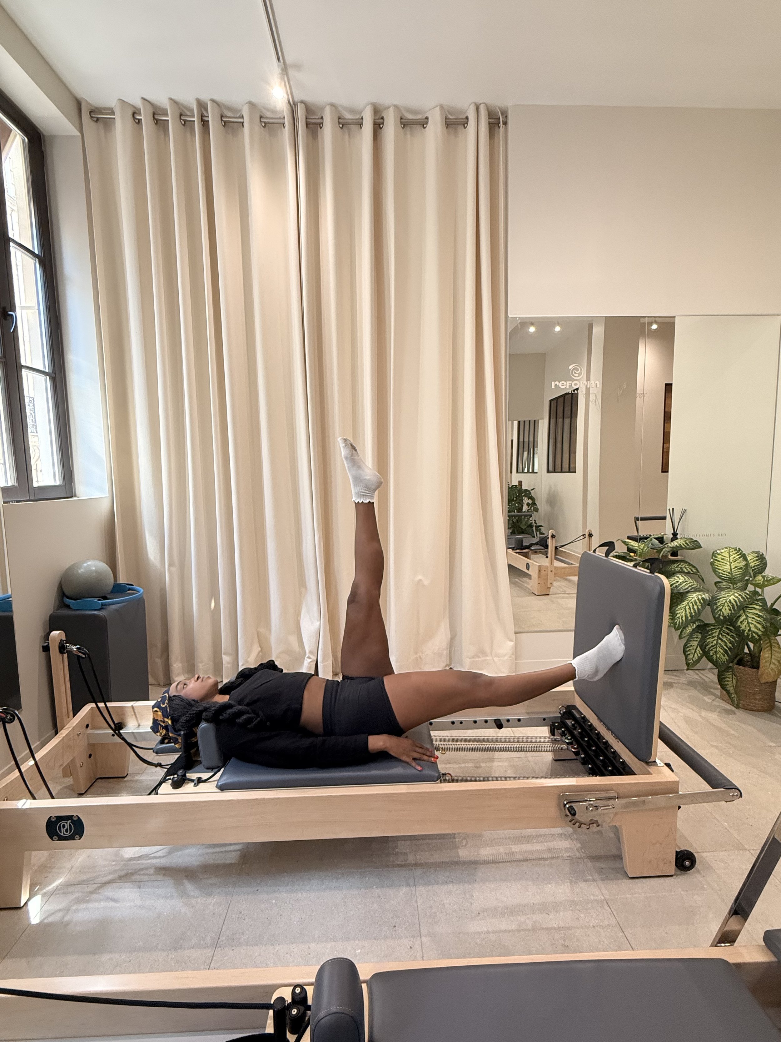 Une femme effectuant un exercice de Pilates sur une machine reformer dans une studio bien éclairée avec des plantes et des rideaux beige.