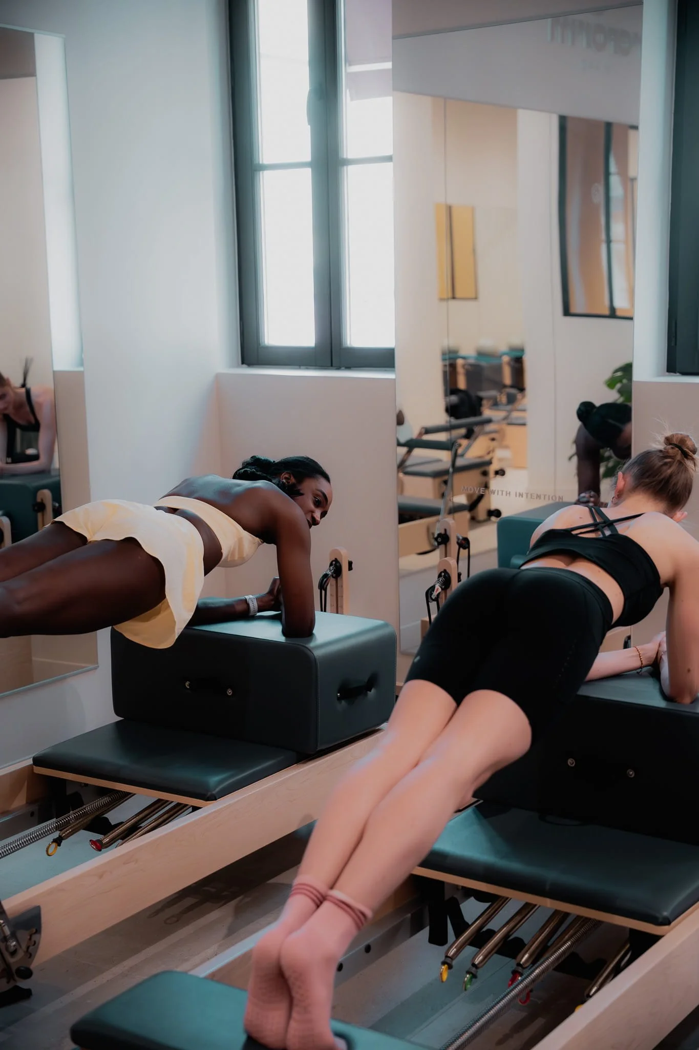 Groupe de personnes faisant du pilates ou de l'exercice dans un studio bien éclairé avec grandes fenêtres. Il y a un miroir sur le mur et des équipements d'exercice.