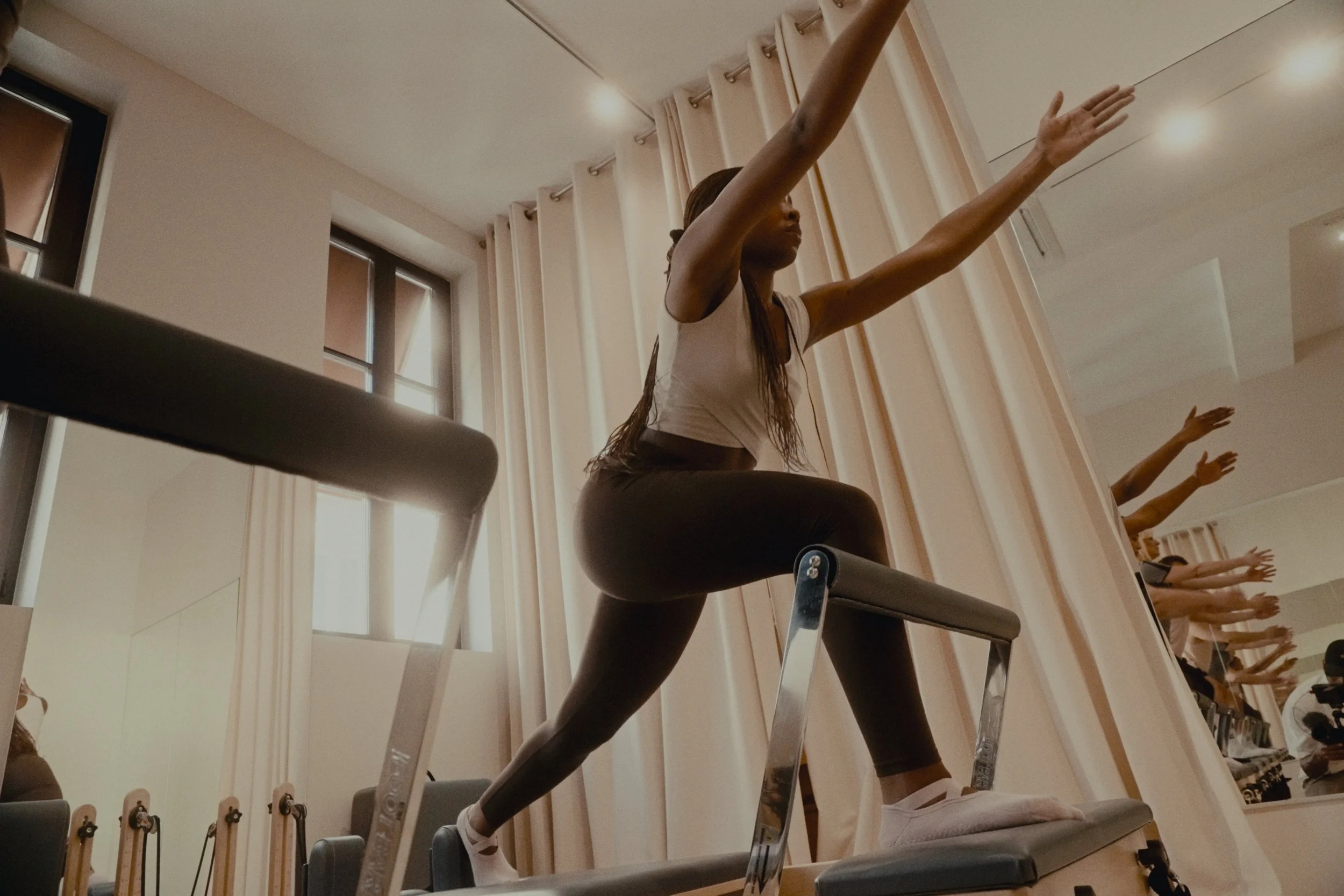 Femme pratiquant le pilates sur un reformer, en position de squat avec les bras levés dans une salle lumineuse avec grandes fenêtres.