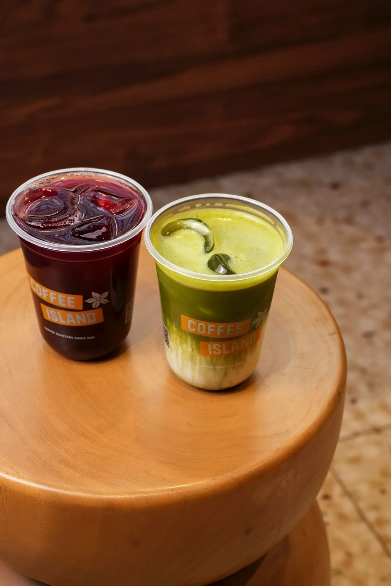 Deux boissons colorées , matcha et latte ,  dans des verres en plastique portant un logo 'Coffee Island' sont placées sur une table en bois rond. L'une est rouge avec des glaçons, l'autre est vert avec des morceaux de fruits.
