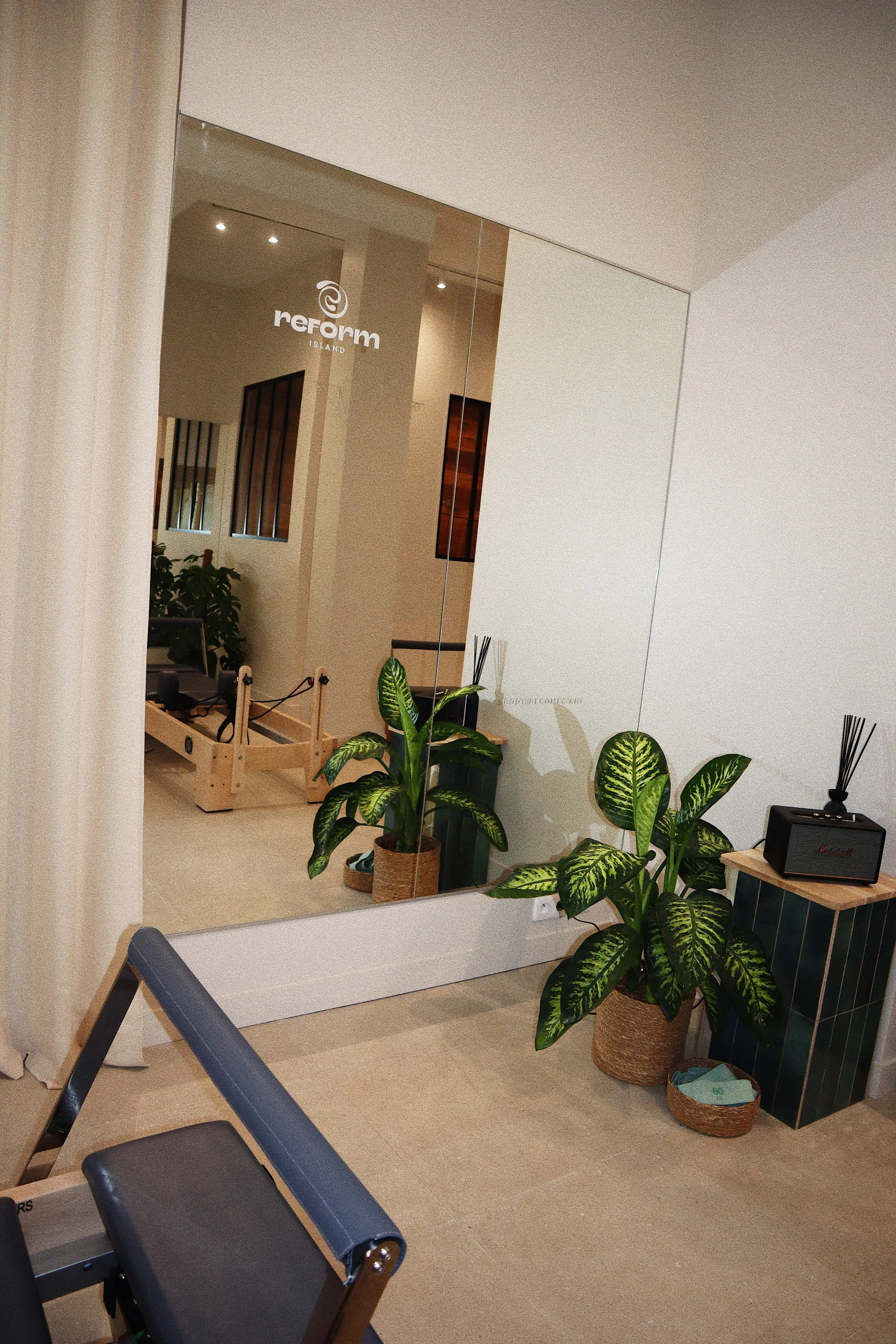 Intérieur d'un espace de soins ou de détente avec des plantes vertes en pot, un grand miroir et une table avec un diffuseur d'huiles essentielles. On aperçoit aussi des machines de pilâtes reformer . La décoration est moderne et épurée.