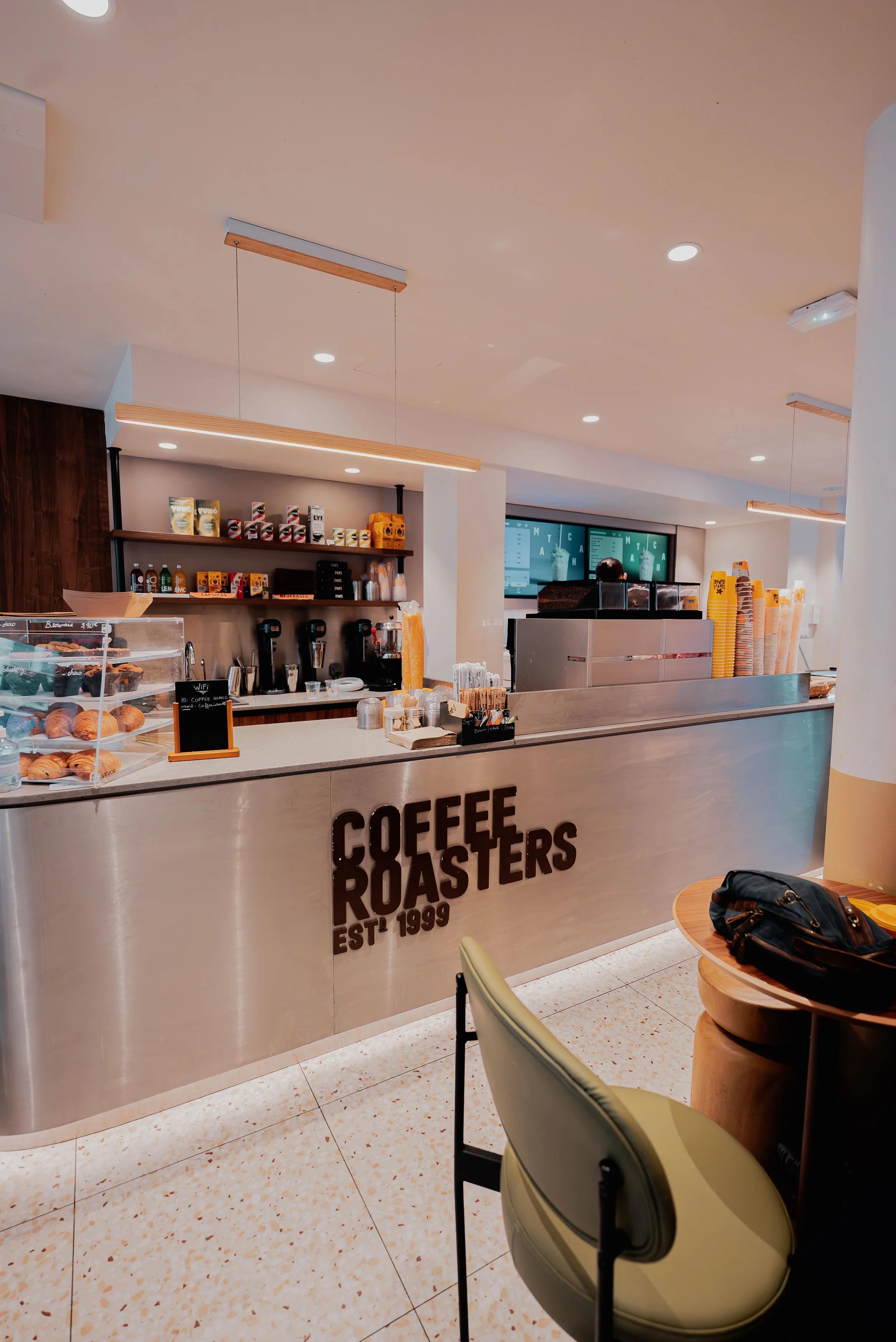 Intérieur d'un café avec un comptoir en inox et affichage digital, des étagères avec des produits en paquets, une table en bois avec un sac à main, une chaise verte, et des lumières suspendues au plafond.