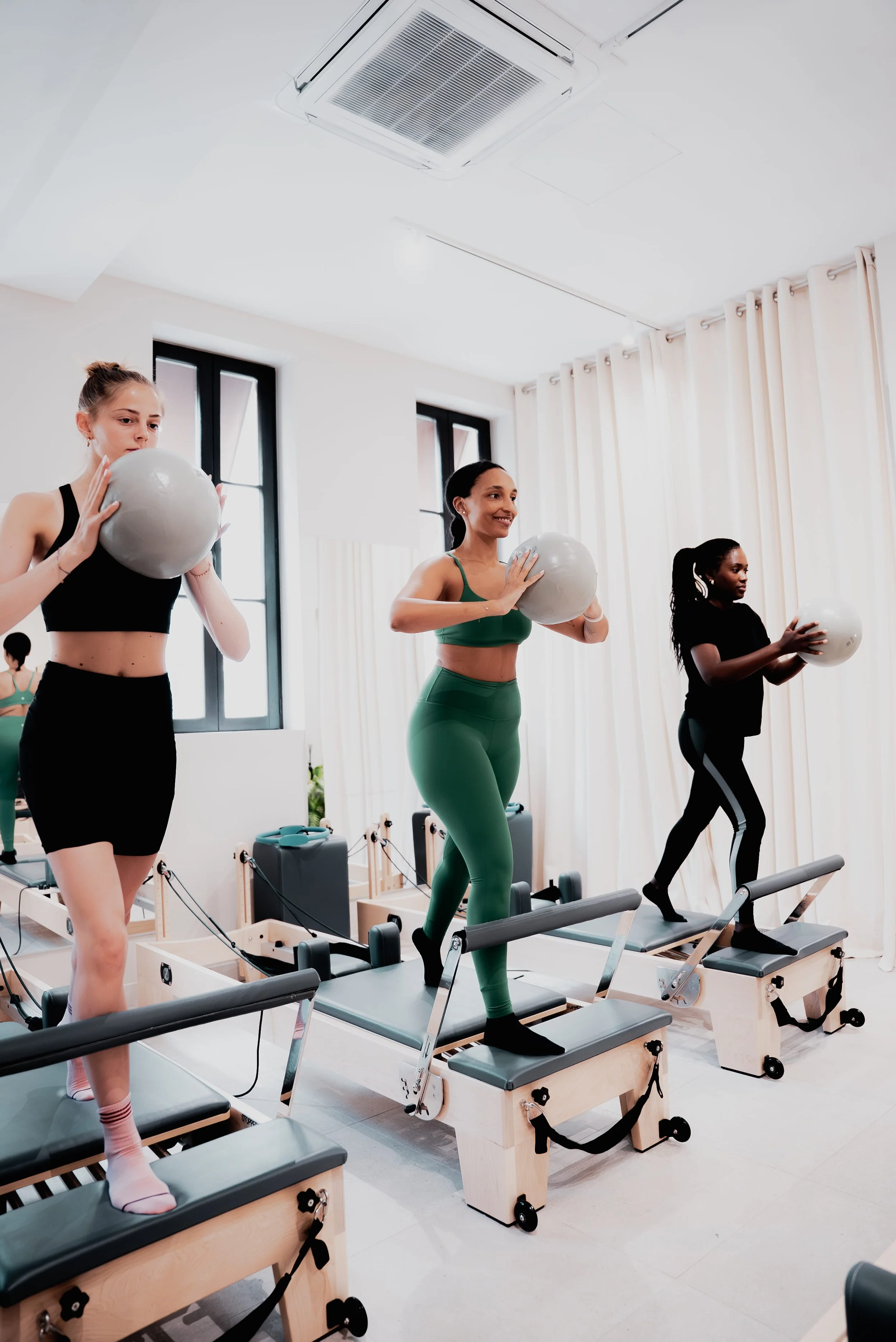 Trois femmes participent à une séance d'exercice sur des machines de fitness dans une salle lumineuse avec grands fenêtres et rideaux blancs.