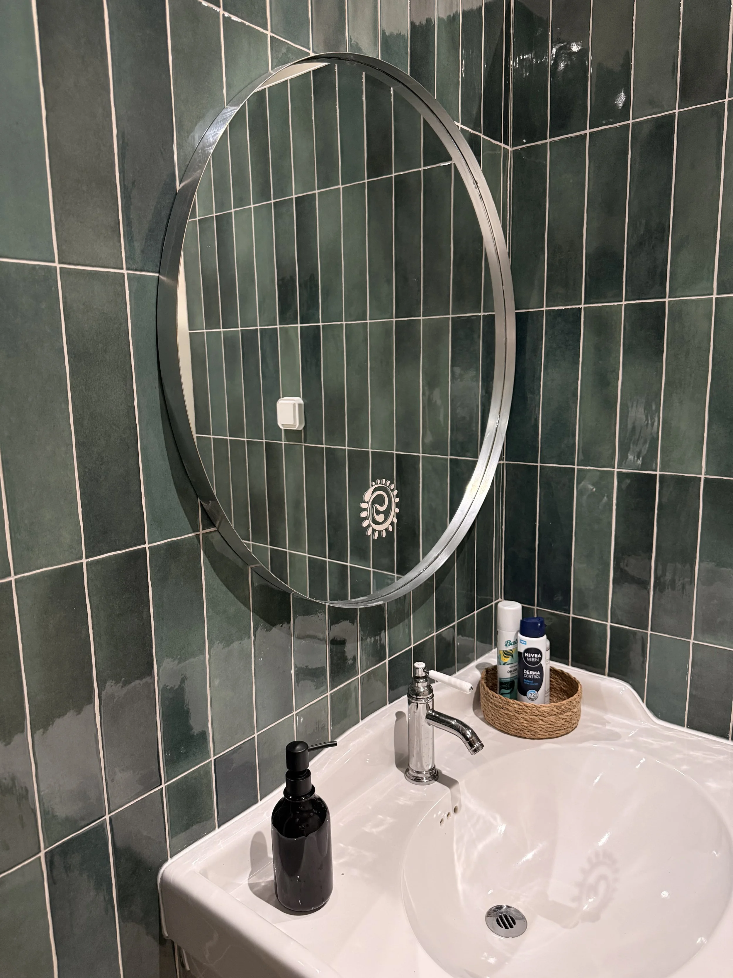 Un miroir rond au-dessus d'un lavabo blanc dans une salle de bain. Le mur derrière est recouvert de carreaux verts foncés. Sur le lavabo, il y a un panier contenant des produits de soins, notamment une bouteille de Nivea Men, une crème Derma Control et une autre crème dans un flacon blanc. Il y a aussi un savon liquide noir et un miroir rond avec un cadre métallique.