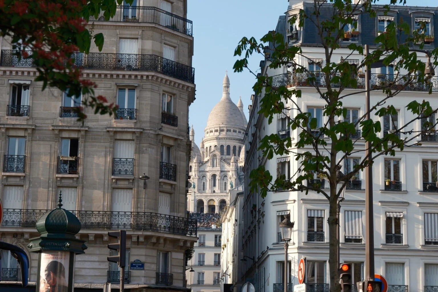 Vue urbaine de Paris avec des immeubles haussmanniens et la basilique du Sacré-Cœur en arrière-plan, sous un ciel clair.
