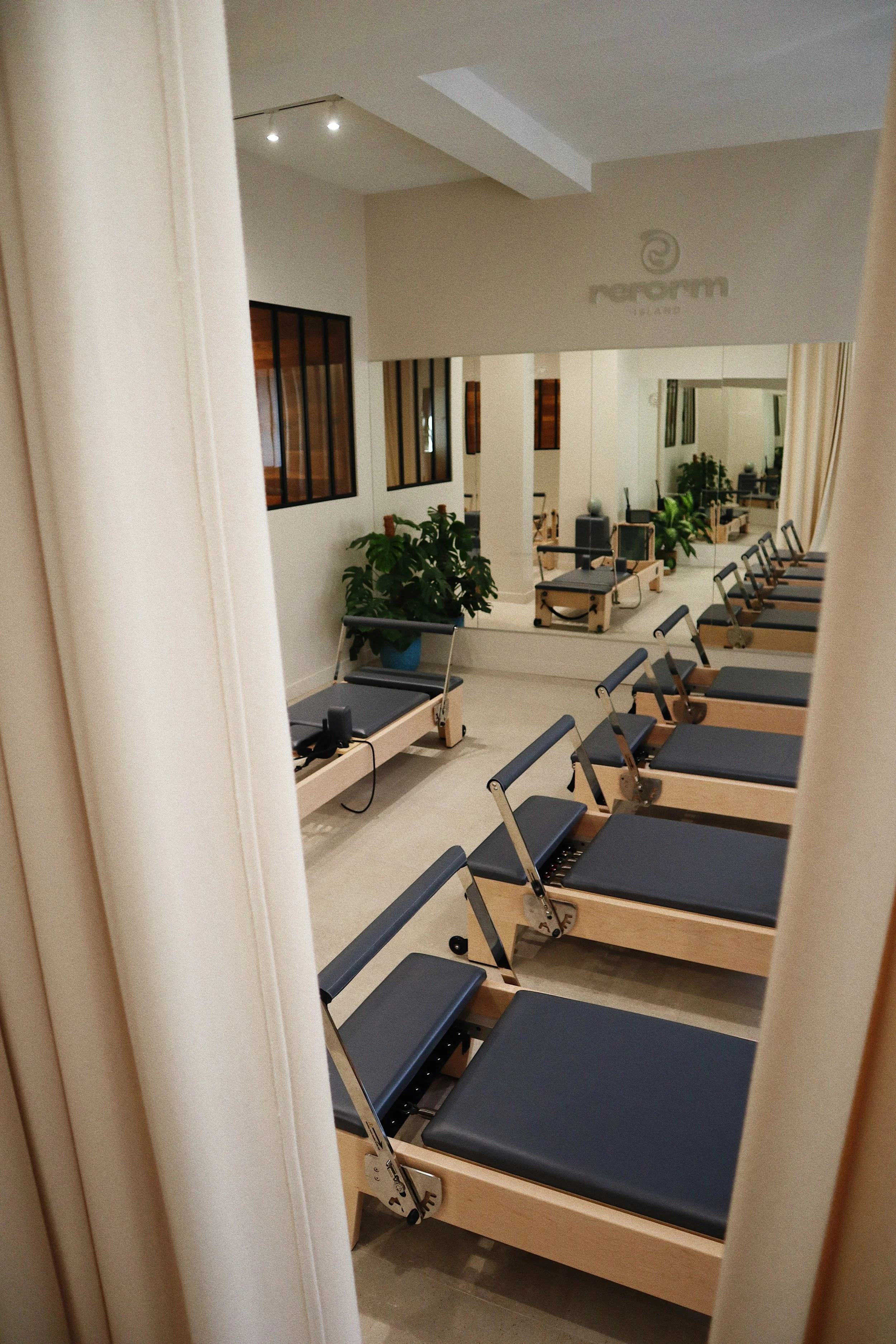 Salle de Pilates avec plusieurs machines de reformer alignées, murs blancs, miroirs et plantes vertes, ambiance moderne et épurée.