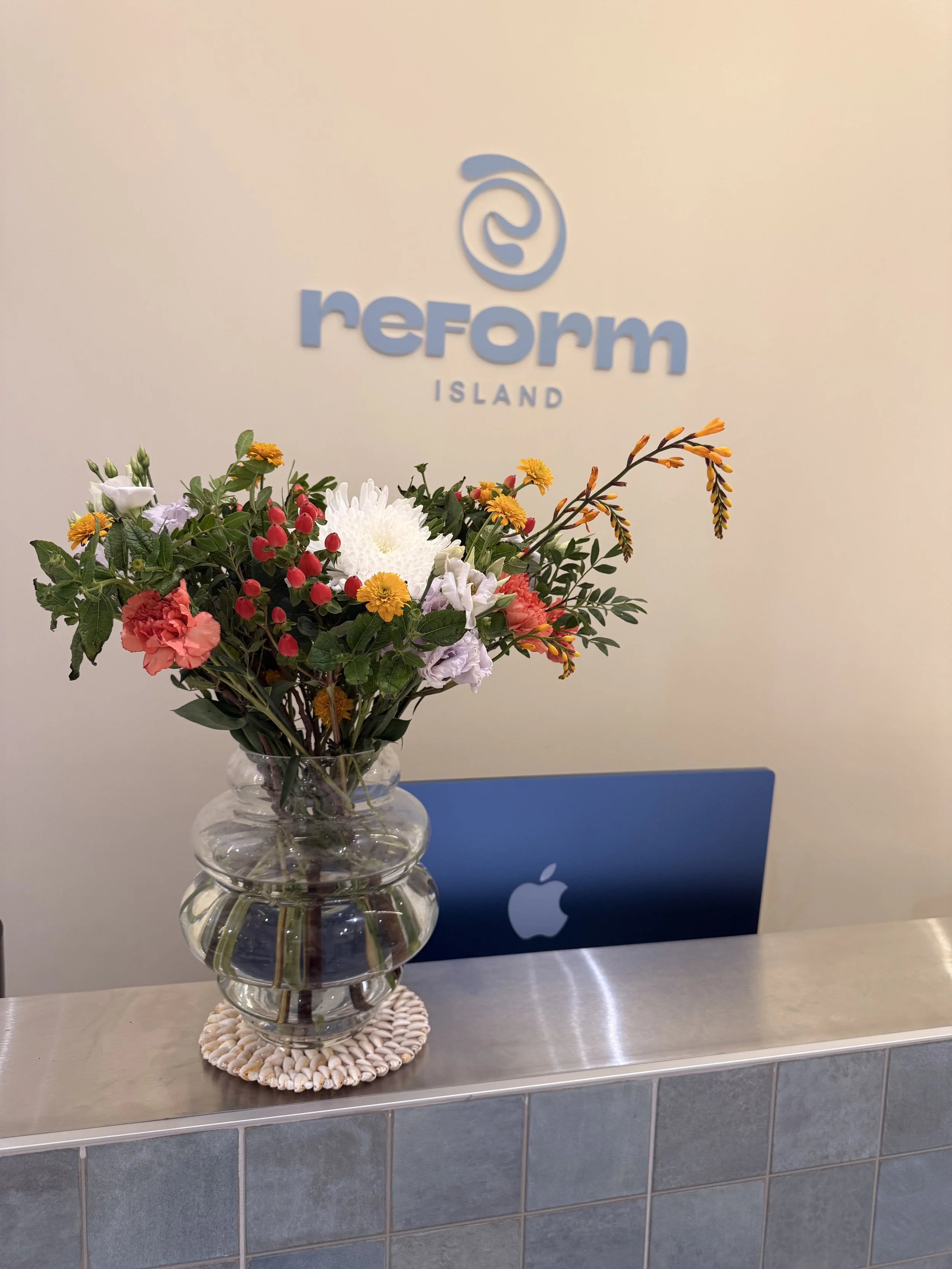 Un vase en verre transparent rempli de fleurs colorées, placé sur un tapis en coquillages, avec un logo Apple en arrière-plan sur un mur blanc avec le mot 'reform' et 'ISLAND' en dessous.