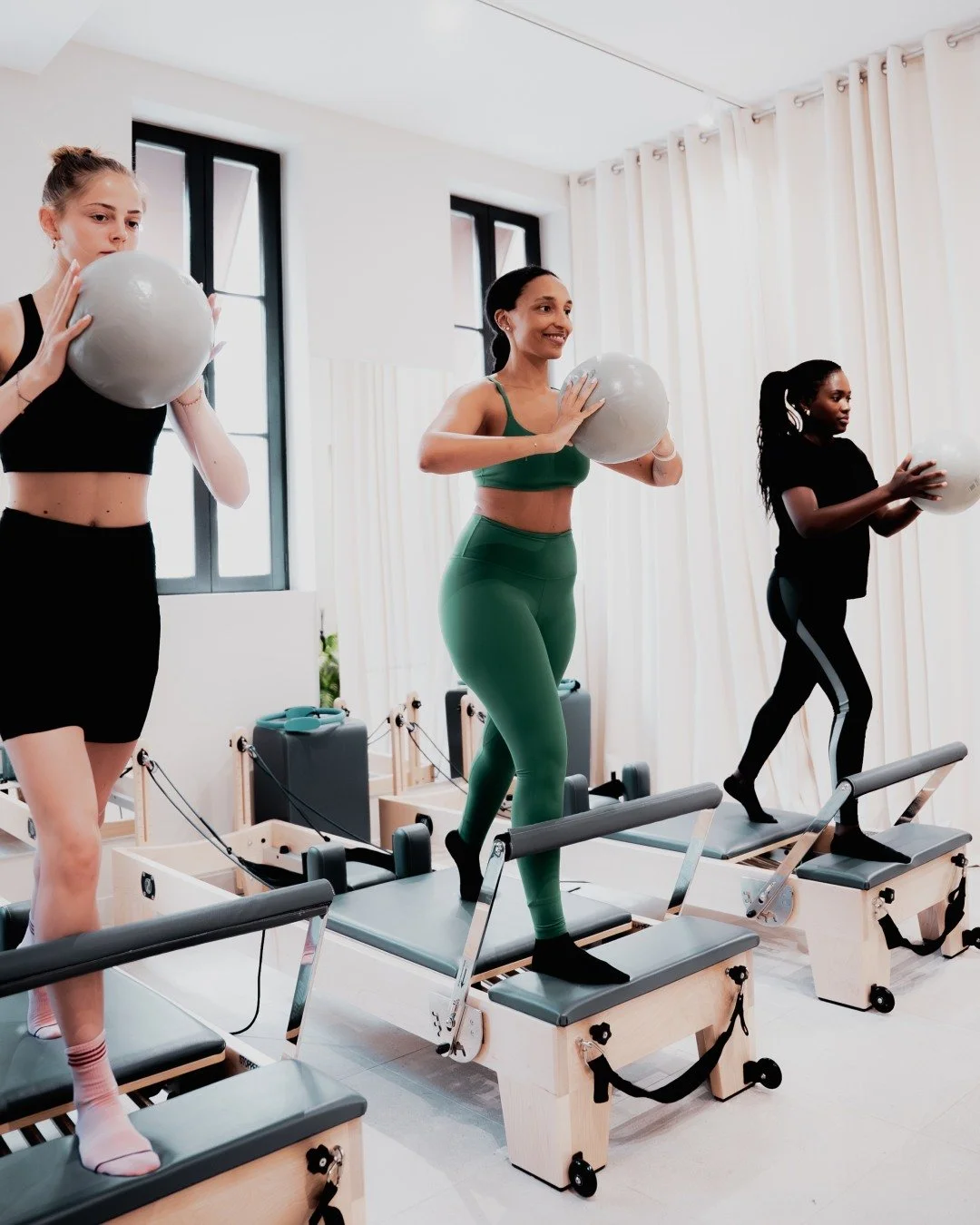 Trois femmes faisant de l'exercice sur des Reformer Pilates avec des ballons dans une salle lumineuse avec fenêtres.