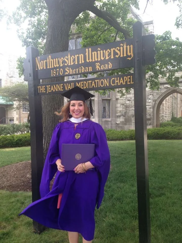 A woman in a purple graduation gown and cap holding a diploma, standing in front of a campus sign that reads 'Northwestern University 1870 Sheridan Road', near The Jeanne Vail Meditation Chapel, on a grassy area with bushes and trees.