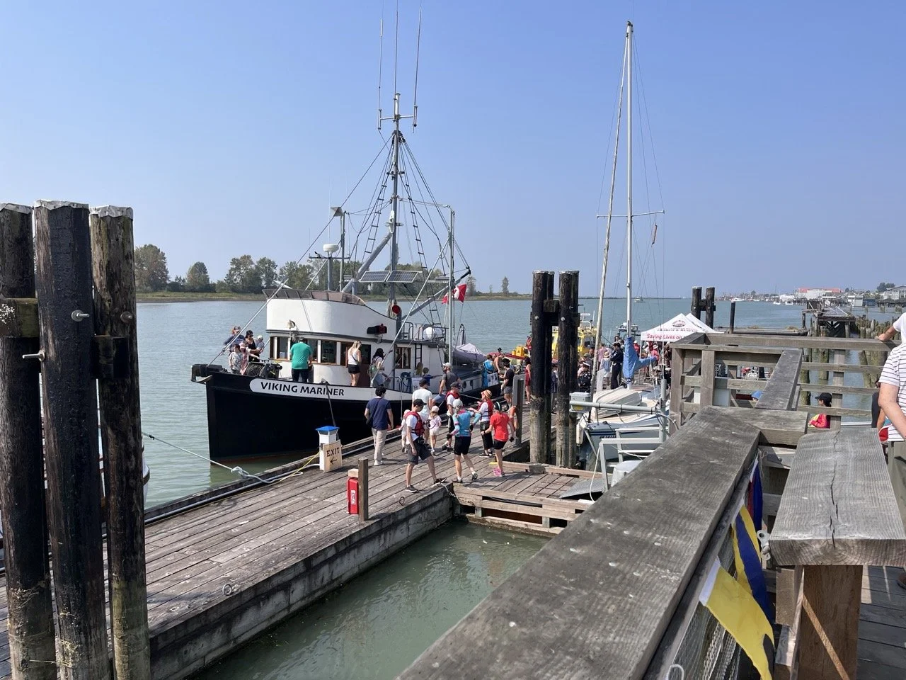 People boarding a boat at a marina dock on a sunny day, with clear blue sky and calm water, surrounded by wooden pier and posts.