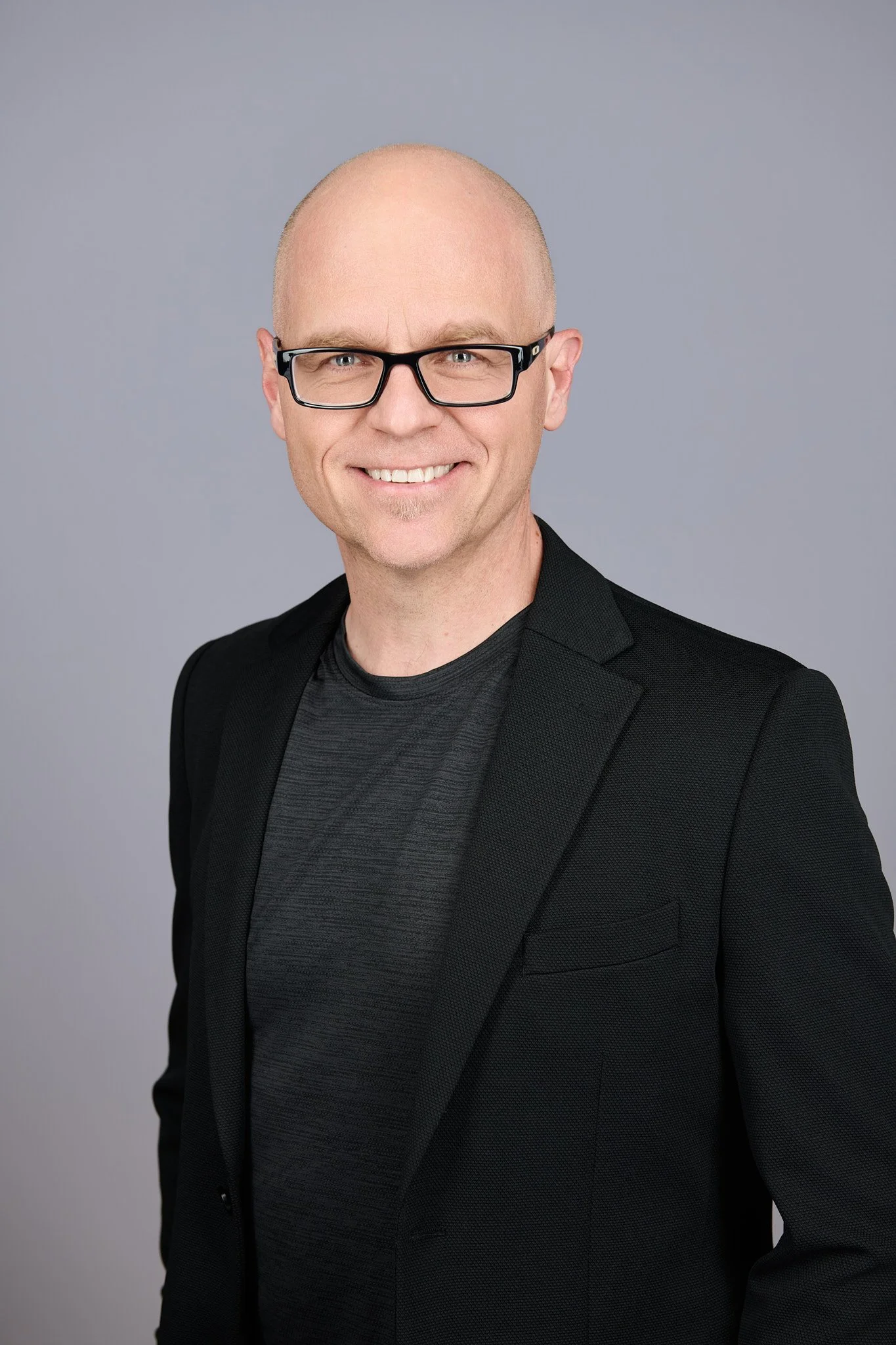 Portrait of Tamas smiling, wearing a black blazer over a dark gray shirt, against a plain gray background.