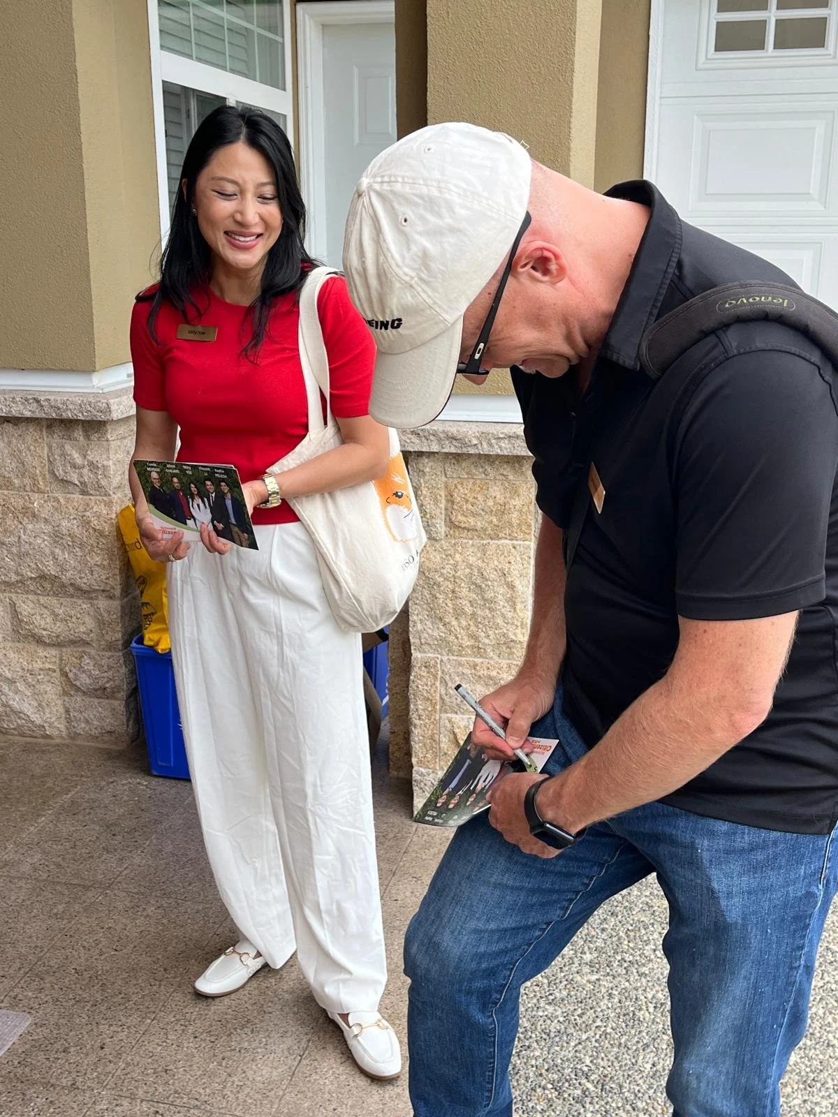 A woman in a red shirt and white pants holding a flyer, talking to a man in a black shirt and jeans who is signing an autograph.