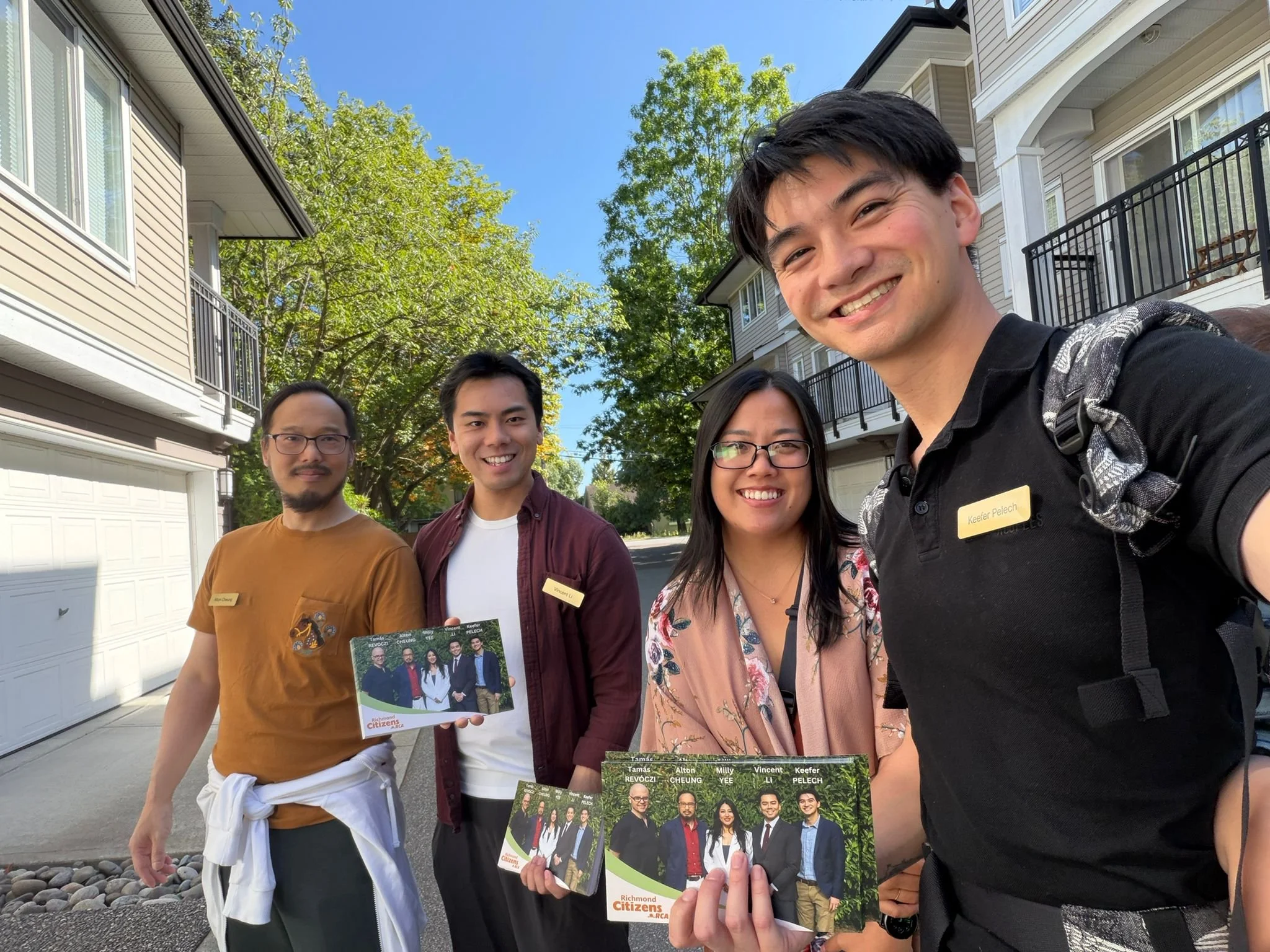 A group of five people standing outdoors on a sunny day, holding brochures and smiling for a selfie. The group is diverse and looks happy, with a background of residential houses and trees.