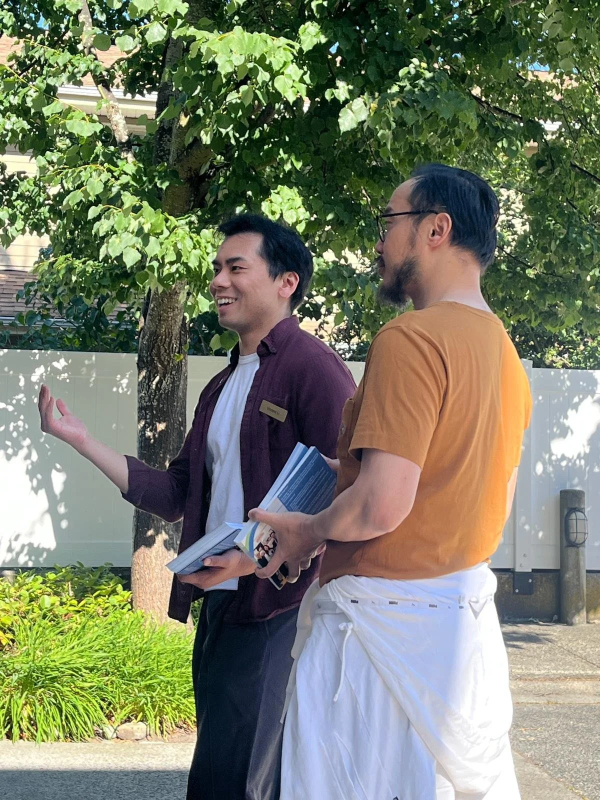 Two men conversing outdoors beside a tree and white fence. One man, smiling, holds flyers or brochures. The other, with glasses and a beard, listens.