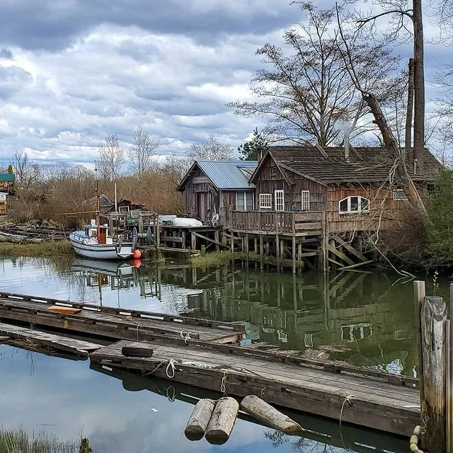 A rustic wooden boathouse and dock beside a small waterway with a boat moored to the dock. The scene has cloudy skies and leafless trees, suggesting late fall or winter.