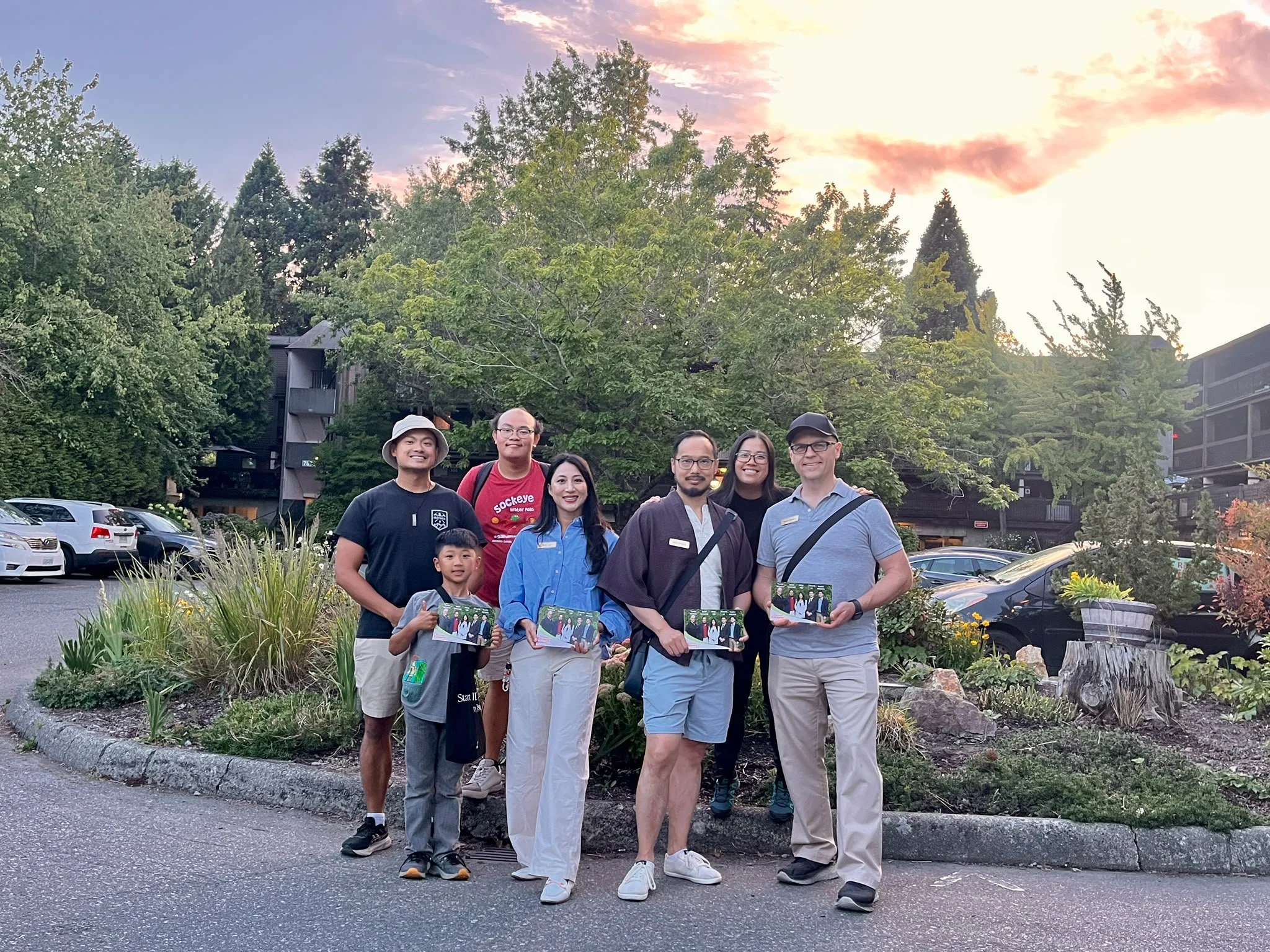 A group of seven people, including children and adults, standing outdoors on a sidewalk with lush green trees and a sunset sky in the background, holding photographs or flyers.