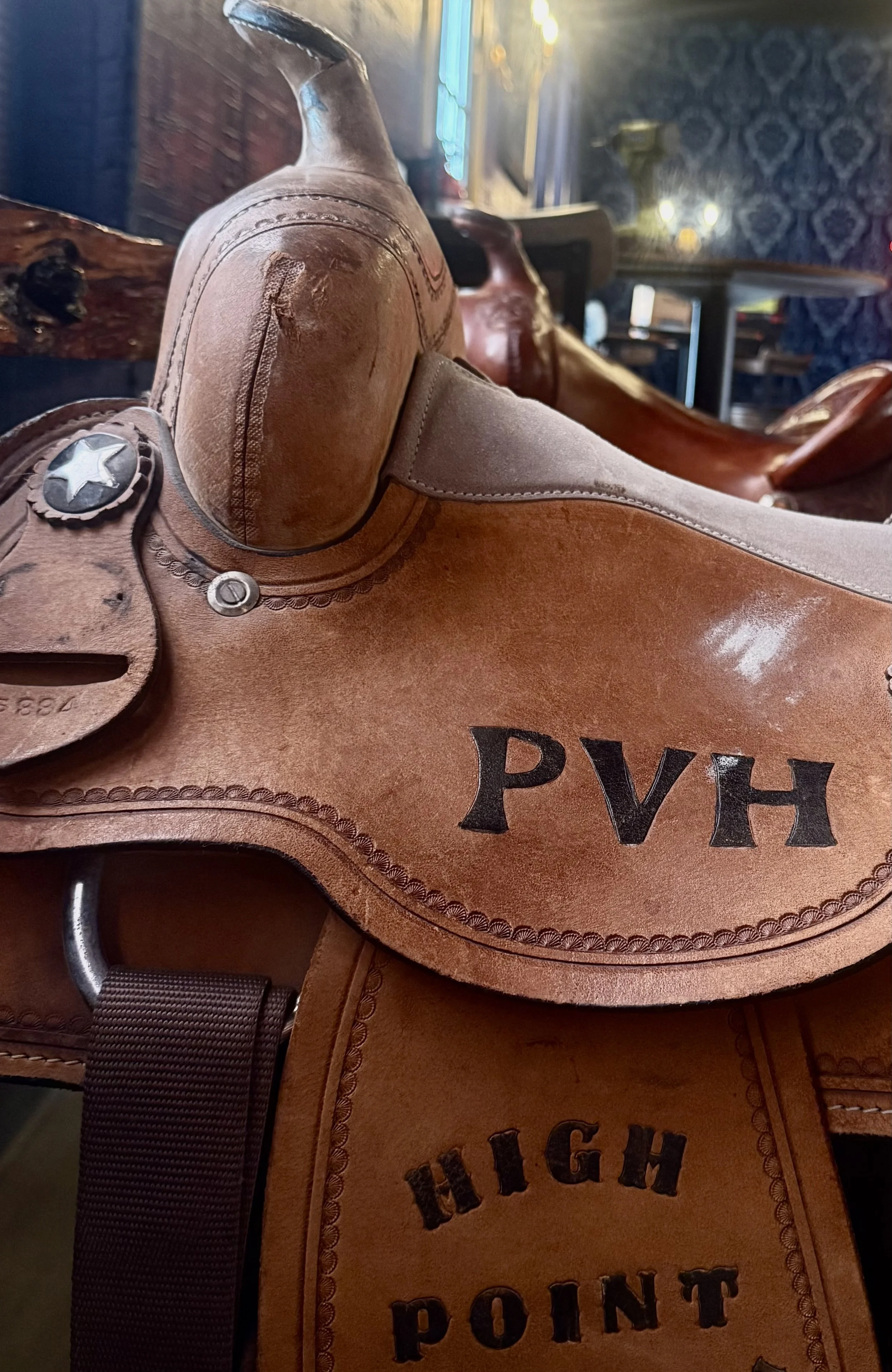Close-up of a cowboy saddle with leather details and decorative stitching, set against a blurred background of a dimly lit bar or restaurant with patterned wallpaper and tables.