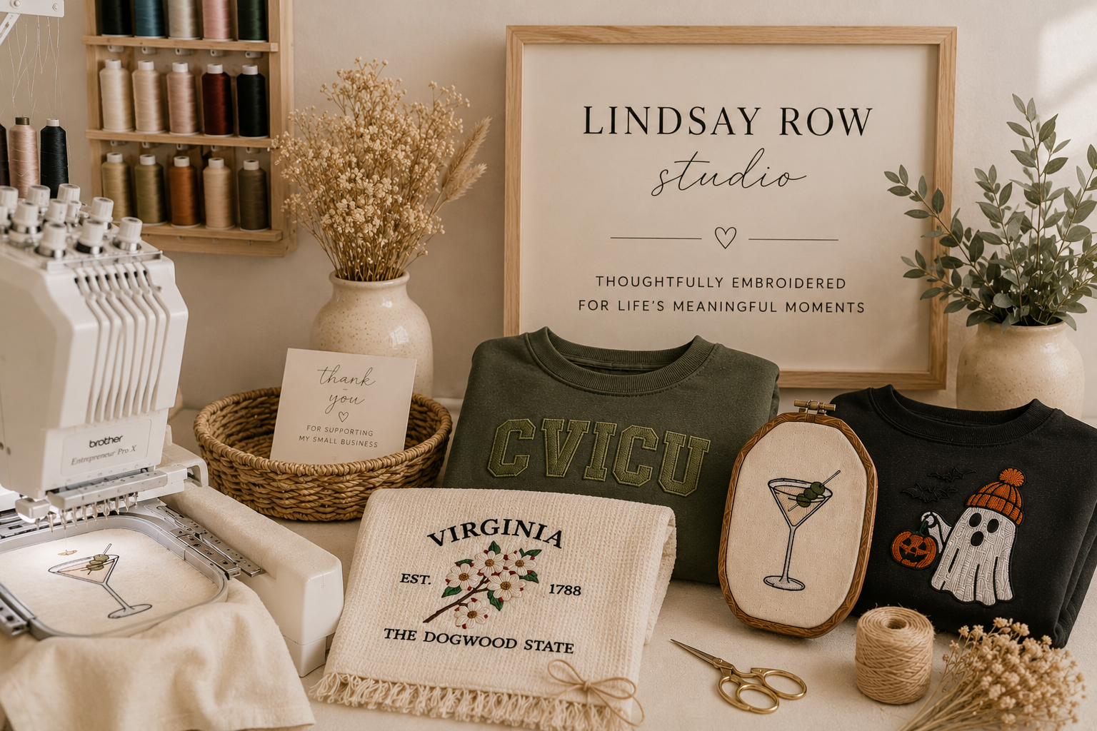 A display of embroidery projects and supplies on a table, including embroidered towels, shirts, framed artwork, spools of thread on a wall, silk flowers in a vase, embroidery scissors, a small basket with a thank you note, and embroidery hoop with a martini glass design.