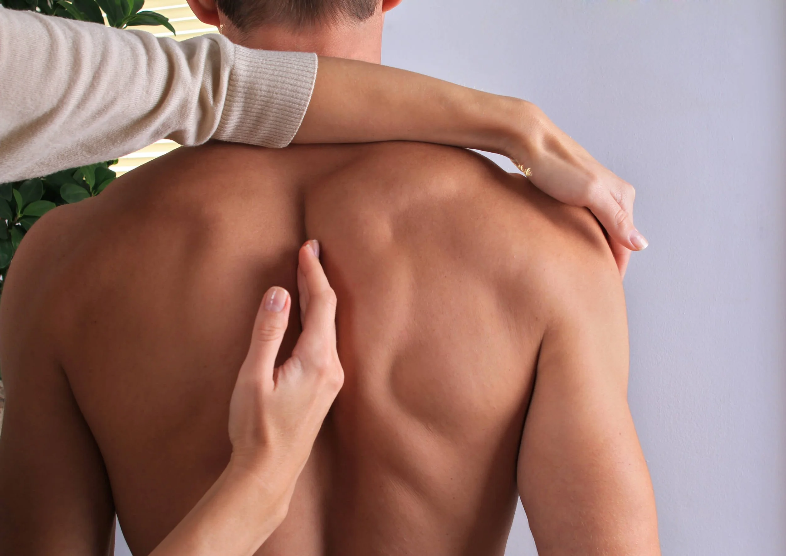 A person with light skin and short brown hair is receiving a massage or physical therapy from a second person with light skin, whose hand is placed on the person's back. The background includes a light-colored wall and some greenery.