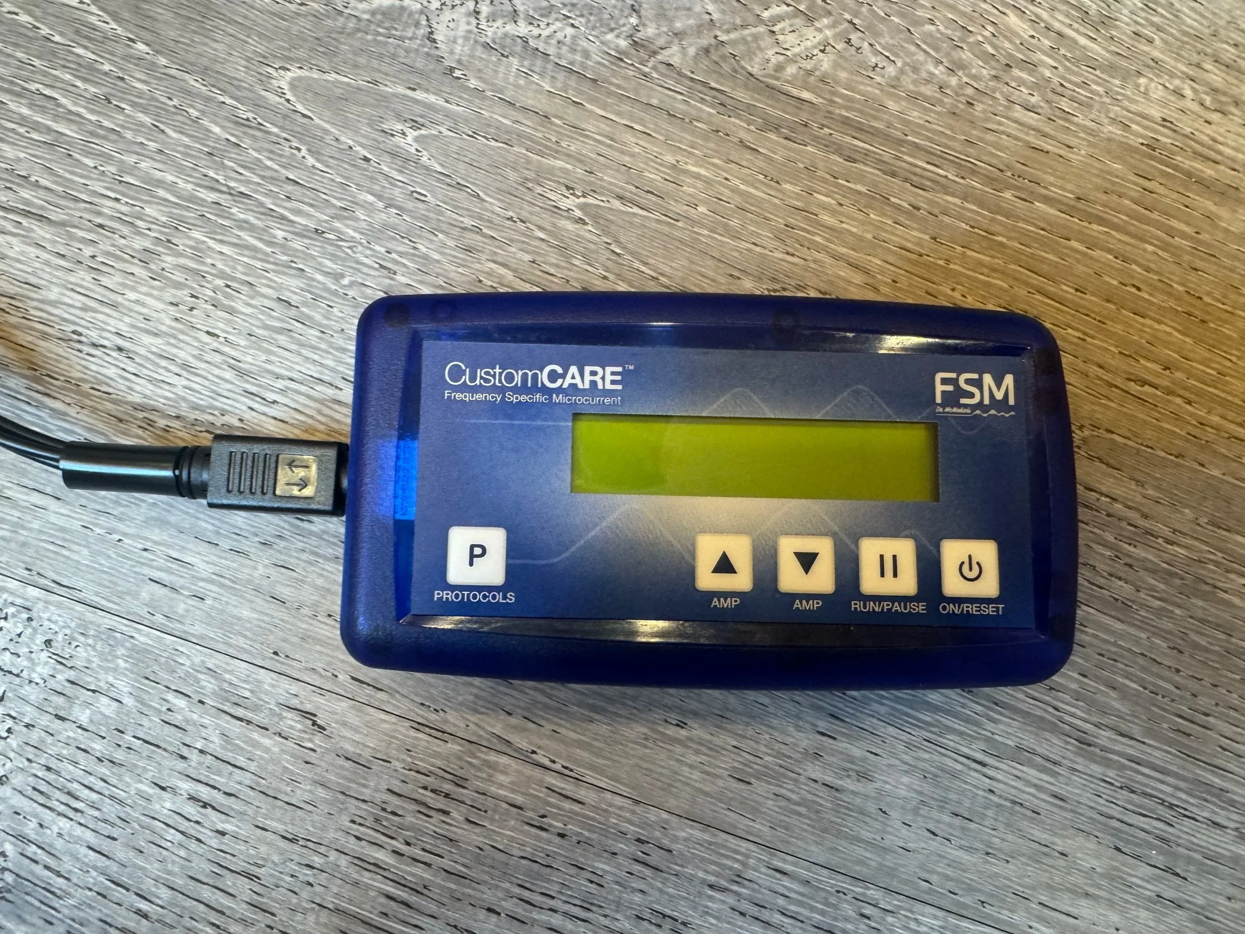 Close-up of a blue medical device labeled 'CustomCARE' with a digital screen and control buttons, connected to a black cord on a wooden surface.