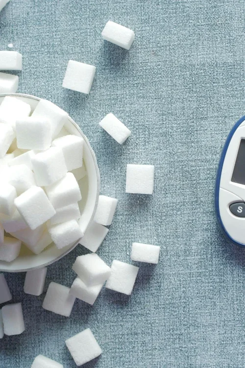A bowl of sugar cubes spilled onto a blue textured surface next to a blood glucose meter.