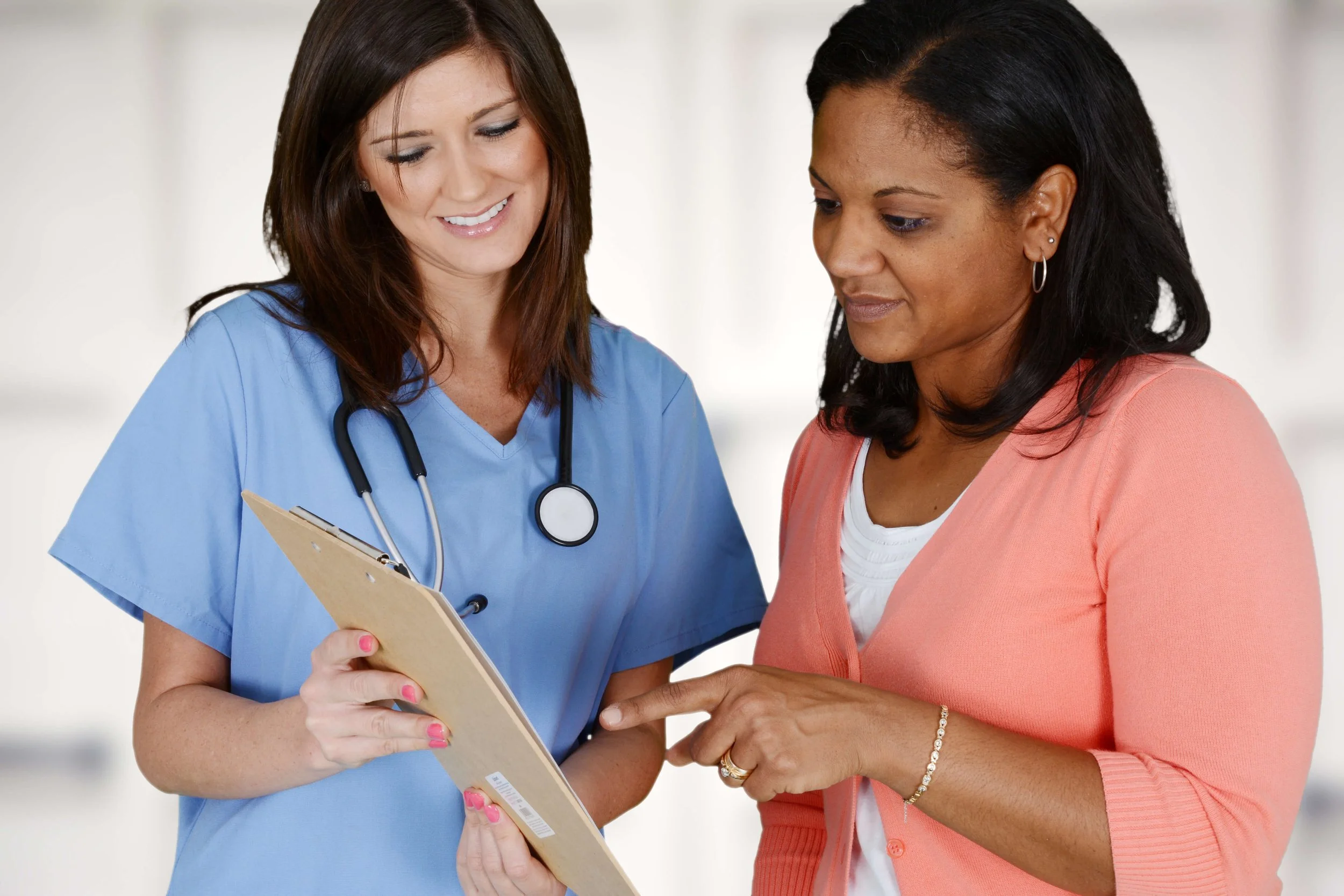 A healthcare professional in blue scrubs and a stethoscope around her neck shows a clipboard to a patient in pink clothing. They appear to be discussing medical information.
