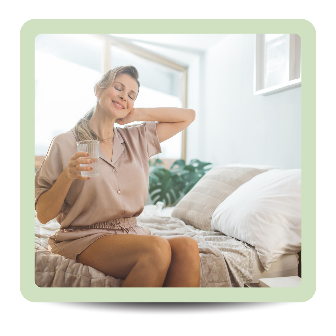 A woman sitting on a bed with a glass of water, smiling and stretching in a bright bedroom.
