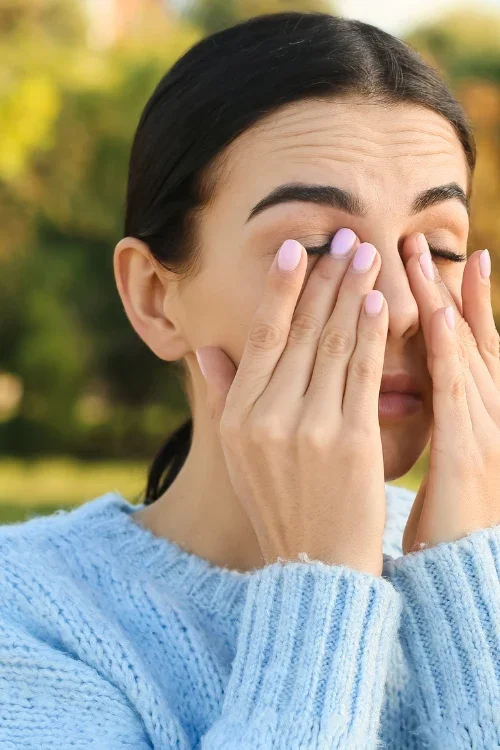 A woman with dark hair and light skin wearing a light blue sweater is outdoors, covering her face with her hands, possibly rubbing her eyes or sneezing, with a blurred background of trees and greenery.