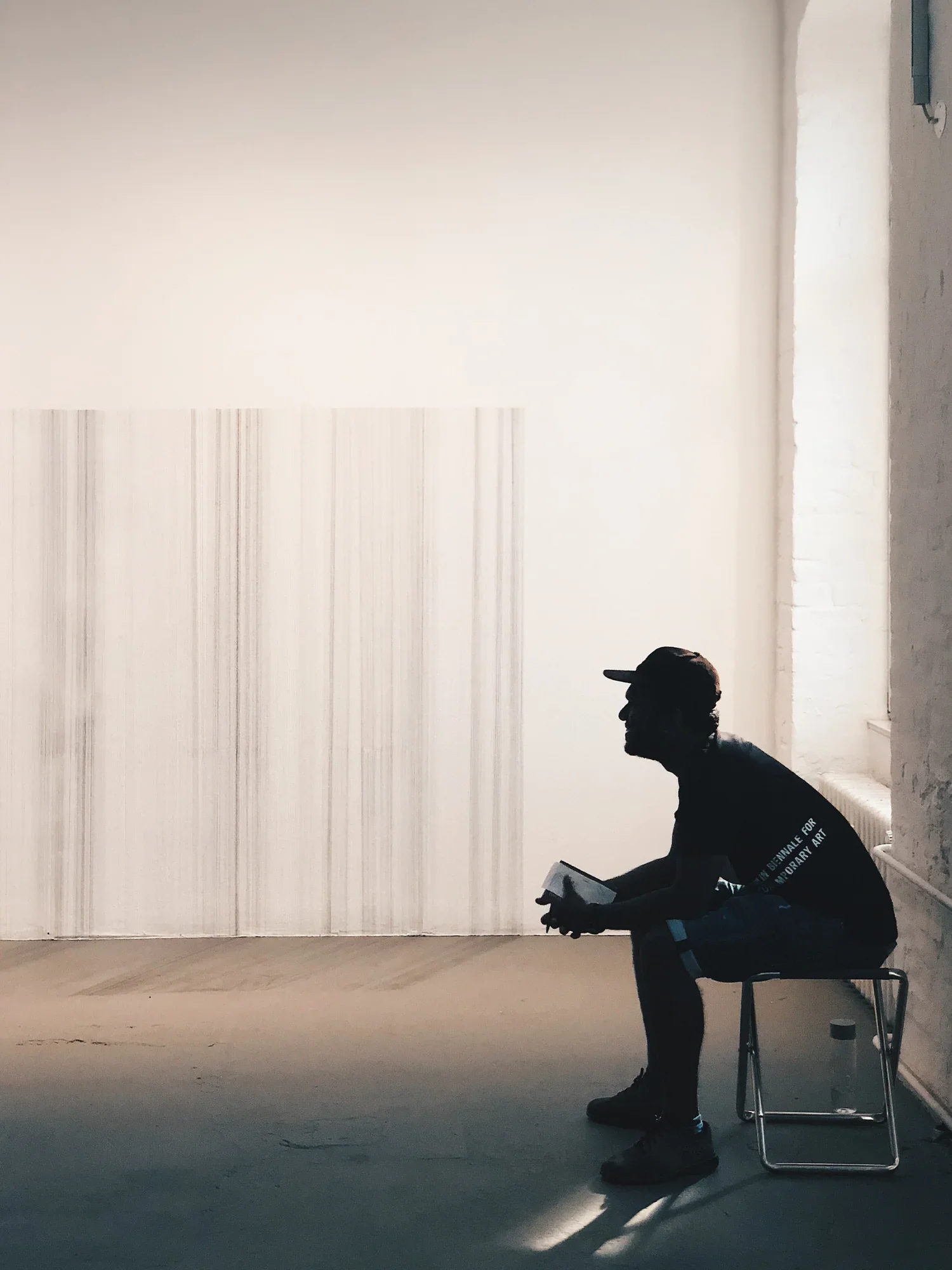 Silhouette of a person sitting on a chair, holding a book, in a minimalistic, white-walled room with a radiant window.