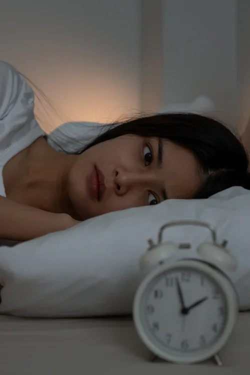 A woman lying in bed looking at an alarm clock with a worried expression.