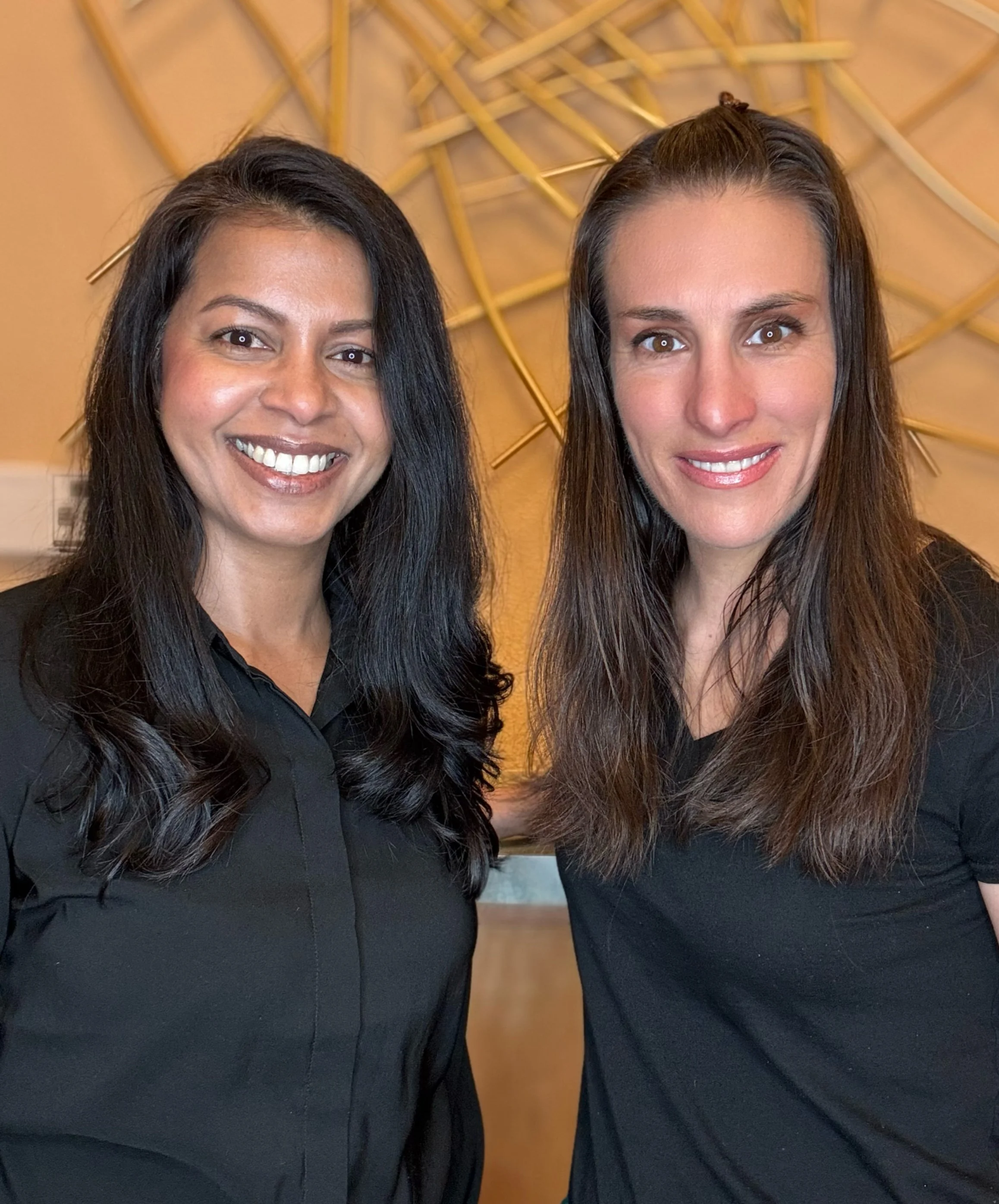 Close-up of two women smiling, standing side by side, with a geometric wall art installation behind them.