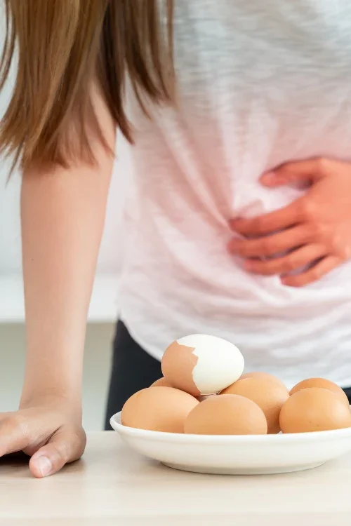 Person with long hair and a white shirt is standing next to a bowl of eggs, one of which is cracked open.