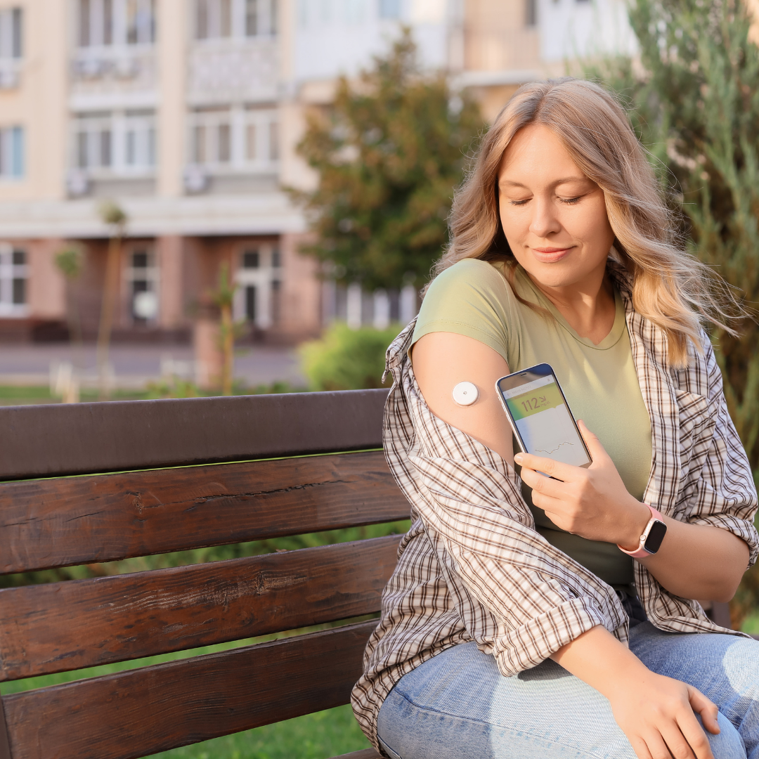 A woman sitting on a park bench outdoors, checking her blood sugar level on her smartphone, with a medical device attached to her arm.