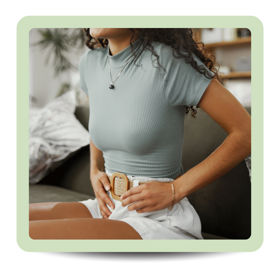 Woman sitting on a couch, wearing a light blue ribbed t-shirt, white shorts, a beige belt, and a necklace, with her hand over her stomach.